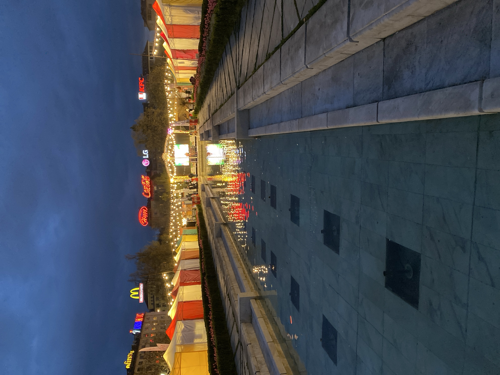 At dusk, a long water feature, possibly a reflecting pool or fountain, occupies the foreground. Its surface reflects the illuminated structures lining the opposite side. These buildings display prominent advertising signage, including brands like "firstfig," "McDonald's," "Happy," "Coca-Cola," "LG," and "KFC". The row of establishments appears to be temporary structures, possibly tents, adorned with string lights. The sky is dark. The location appears to be in Sofia, Bulgaria.
