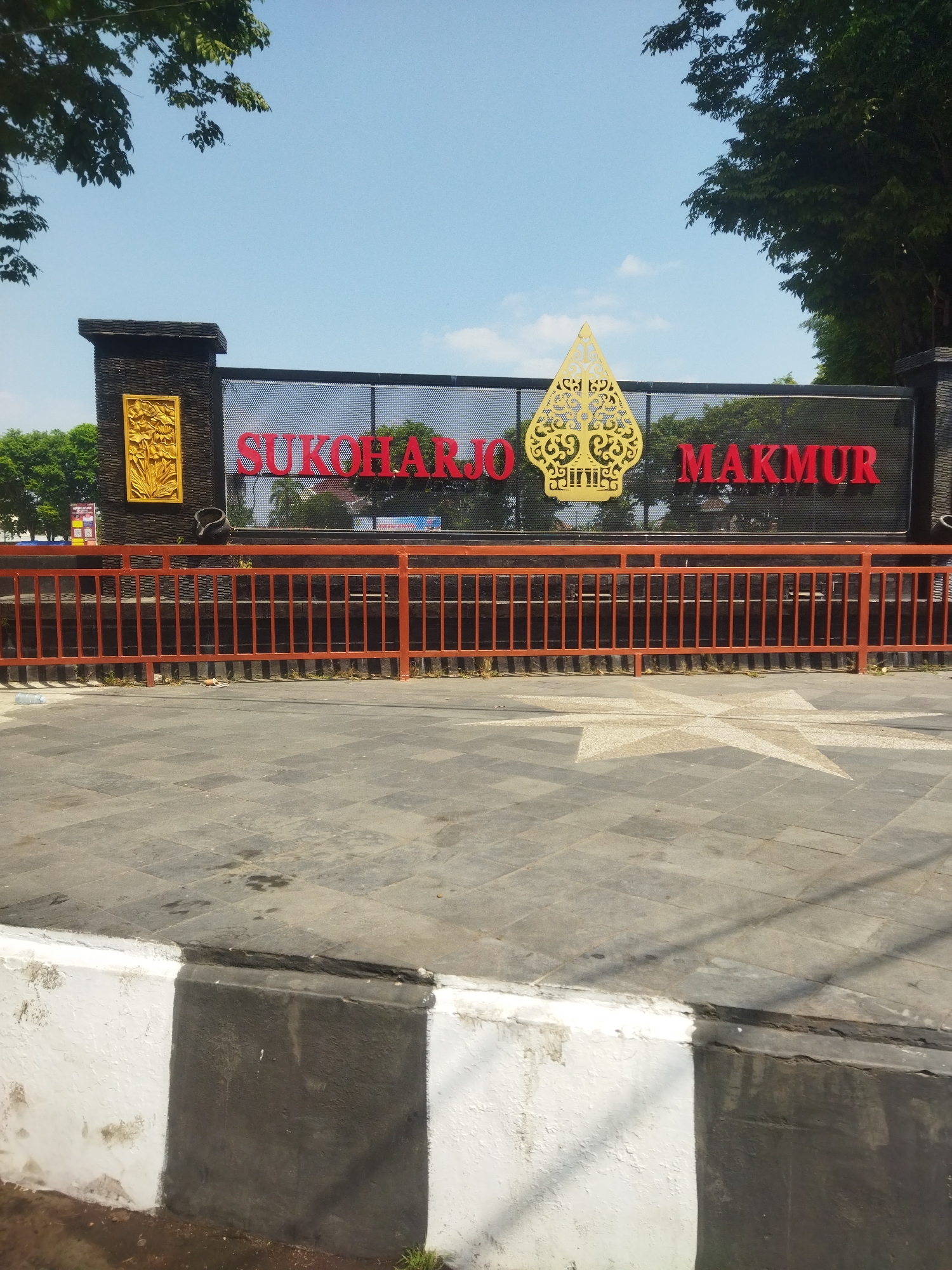 The image shows a sign in Sukoharjo, Indonesia. The sign reads "SUKOHARJO MAKMUR" in large red lettering, with a traditional Javanese Wayang-style emblem in yellow positioned between the two words. The sign is mounted on a black, perforated metal fence. To the left of the sign is a black stone pillar with a gold-colored relief carving of a floral motif. In front of the sign, there is an orange metal railing. The ground in front of the railing is paved with gray square tiles, and a patterned star design is visible on the pavement. A white and black striped concrete barrier is in the foreground, at the bottom of the image. Trees and a blue sky with scattered clouds are visible in the background.