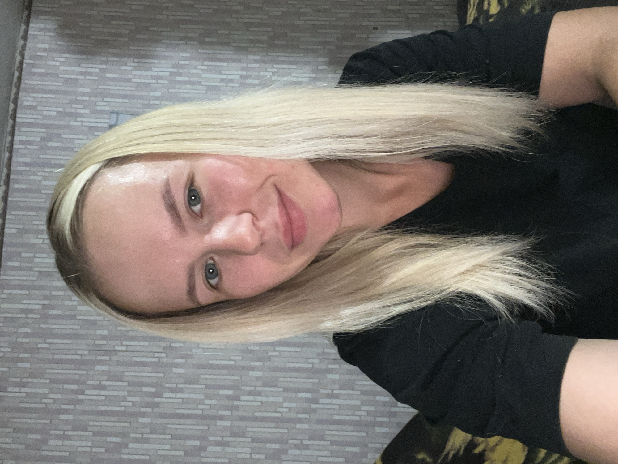 A medium shot shows a young, fair-skinned woman with shoulder-length blonde hair, wearing a black shirt. She has blue eyes and is smiling. The background is a gray, textured wallpaper with a vertical striped pattern. To the left of the woman, part of a couch with a yellow and brown patterned fabric is visible. The lighting appears to be coming from the front, casting soft shadows.