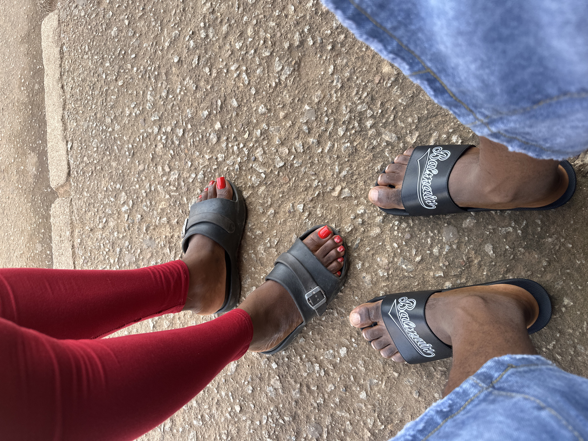 In Bukuru, Nigeria, a low-angle shot captures the feet of two individuals standing on a textured, gravelly surface. One person, wearing vibrant red leggings, has their feet adorned with black slide sandals. Their toenails are painted a bold red. The other individual is dressed in blue denim jeans and black slide sandals featuring a white logo. The scene is rendered in natural lighting, creating a matte finish to the ground and clothing.