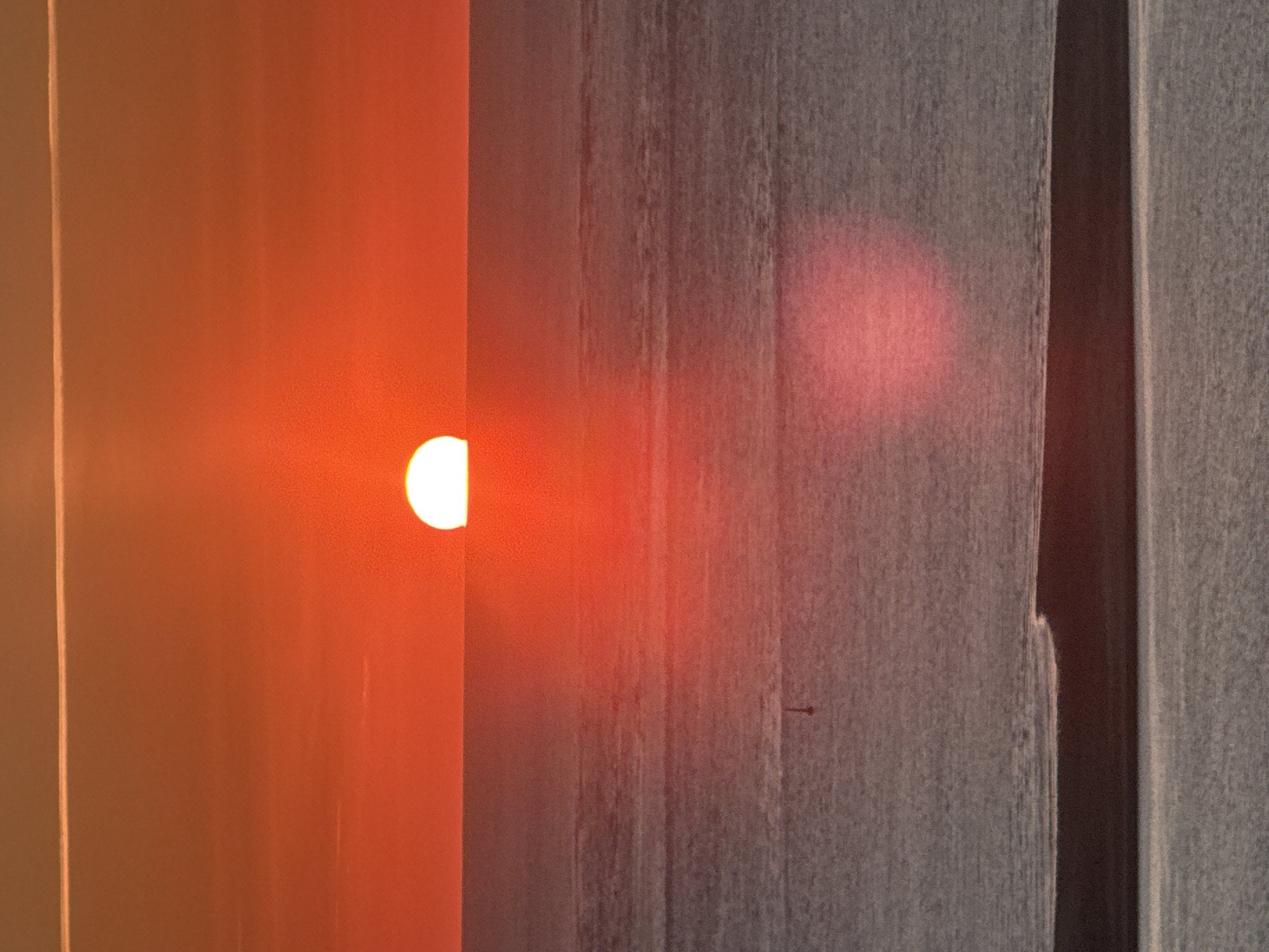 The image captures a sunset over the ocean, likely near La Teste-de-Buch, France, a coastal town known for its beaches and pine forests. The sun, a bright white orb, is partially submerged below the horizon, casting a warm orange and red glow across the sky and water. The sky is a gradient of vibrant orange and red hues, with a thin, bright white line near the top, possibly a contrail or high-altitude cloud. The ocean surface is calm, with subtle horizontal ripples reflecting the sunset's colors. A small, dark, vertical marker, possibly a buoy, is visible in the middle ground of the water. In the foreground, a dark, horizontal landmass, potentially a sandbar or a low-lying island, separates the body of water from another calmer, reflective surface below. Two large, soft, pinkish-red oval shapes are present in the lower half of the water, likely lens flares or reflections of the sunset. The overall scene evokes a serene and picturesque coastal sunset.