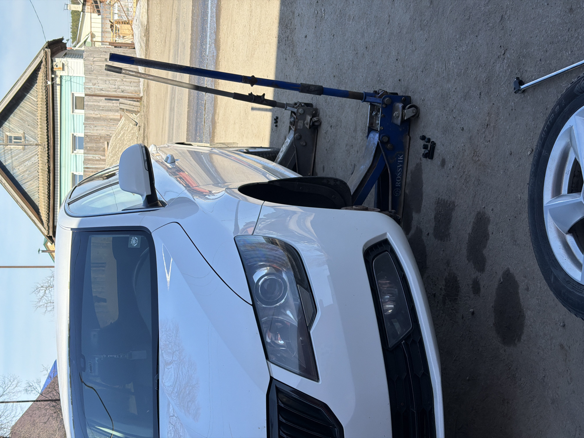 A white car is lifted by two hydraulic jacks on a concrete surface. The car's front driver-side wheel is missing. A silver tire sits on the right side of the frame next to the car. A lug wrench lays next to the tire. A wooden house with blue trim and a brown roof is in the background. The city is Chaikovsky, Russia.