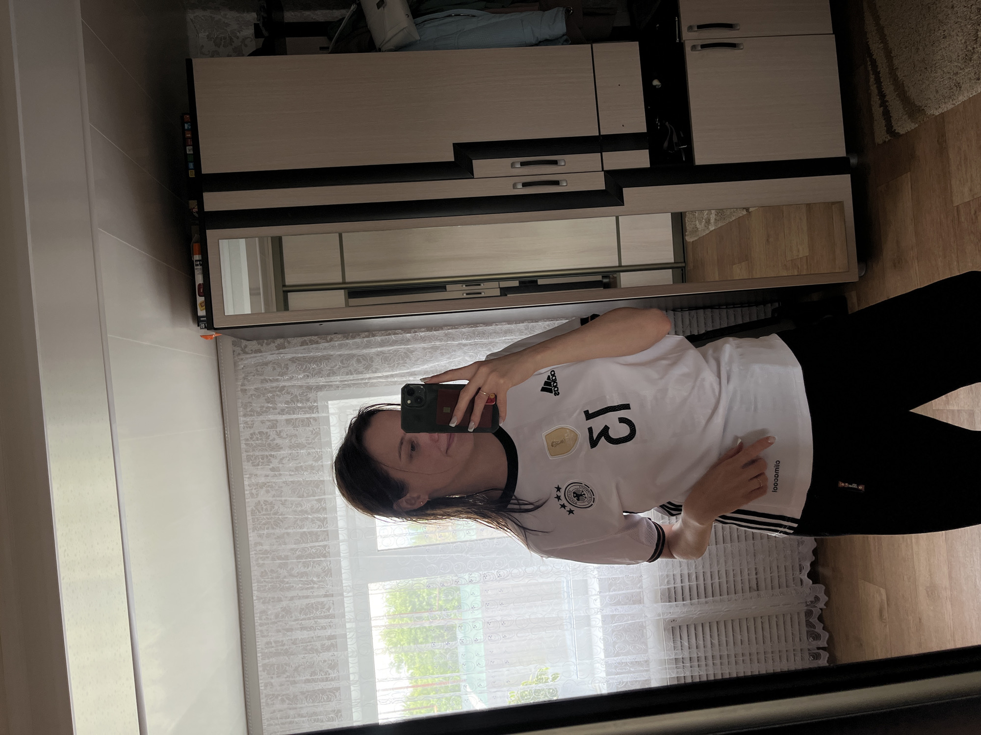 A female individual, estimated to be in her 20s to 30s, is depicted taking a mirror selfie within a residential interior, consistent with a home in a Russian locality such as Novocheremshansk. She stands centrally, directly facing the mirror, holding a smartphone (identified by its camera array, likely an iPhone) in her right hand to capture the image. Her dark, shoulder-length hair appears damp or styled with a wet look. She is wearing a white, short-sleeved athletic shirt featuring black trim on the collar and sleeves. The shirt displays an Adidas logo on the wearer's right chest, a gold FIFA World Cup Winner's badge on the left chest, and the German Football Association (DFB) crest with four stars below it. The number "13" is printed on the front, appearing reversed due to the mirror reflection, as does the word "looooomila" printed on the lower left side of the shirt. She is dressed in black trousers or leggings. A ring is visible on her left ring finger, and her facial expression is a slight, closed-mouth smile. Her left hand rests on her lower abdomen.
The interior features a light-colored, paneled ceiling. A large window, covered by white, sheer lace curtains with a floral pattern, is positioned behind the woman. Through the window, green trees and foliage are visible, along with a partial view of a green roof structure, indicating a natural outdoor environment. To the right of the woman, a multi-section wardrobe or storage unit with light wood-toned panels and prominent dark (black) vertical and horizontal accents stands against a wall. One section of this unit incorporates a full-length mirror, reflecting parts of the room. Another section has two small doors with silver handles. On top of the wardrobe, various items, possibly books or boxes, are stacked. A white handbag and a light blue garment hang from an unseen fixture above the rightmost section of the wardrobe. The floor is covered with light brown wood or laminate panels. A light-colored (beige or cream) shaggy rug is partially visible on the floor near the base of the wardrobe. The wall behind the wardrobe is adorned with light-colored wallpaper featuring a subtle pattern.