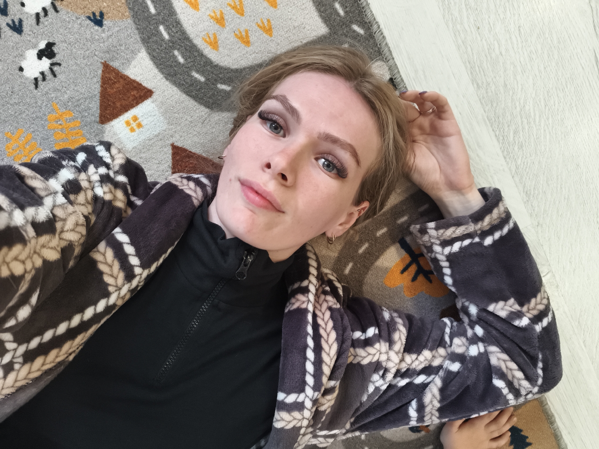 A fair-skinned person with light brown hair is lying on a rug with a road and building design. The person is wearing a black turtleneck shirt and a gray and white patterned robe. The person's right hand is raised near their head, and they are wearing a ring on their ring finger. The person has noticeable eyelashes and fair eyebrows. The rug is on a light-colored wooden floor. The location is Русский Вожой, Russia.