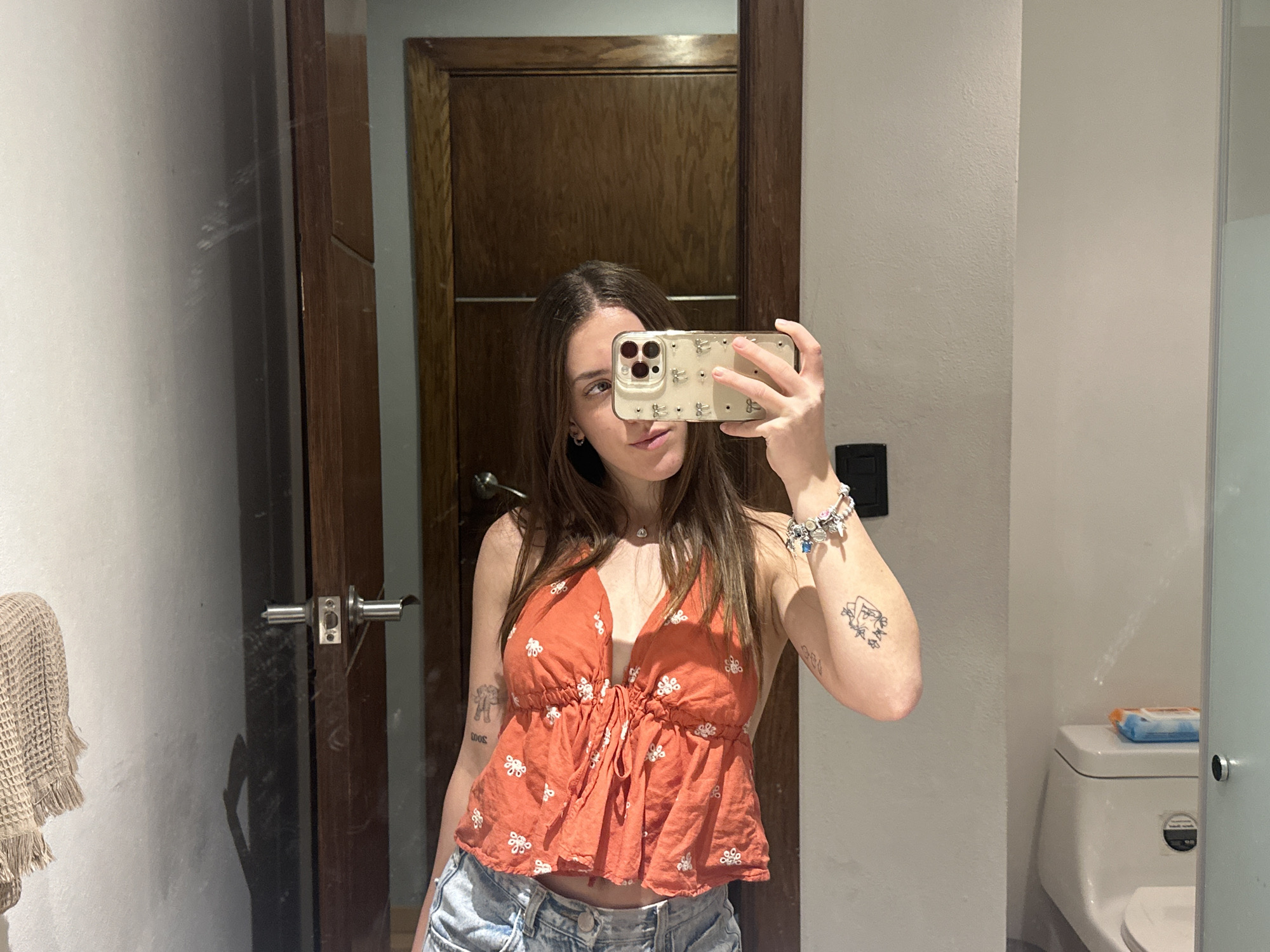 A young woman is taking a selfie in a bathroom. She is wearing a rust-colored floral top and light-wash denim shorts. She holds a phone with a decorative case up to her face, partially obscuring her features. She has a bracelet and a tattoo on her left arm, and another tattoo on her right arm. The background includes a wooden door, white walls, a bathroom mirror, and a toilet with a pack of wipes on top. The location is Huixquilucan de Degollado, Mexico.