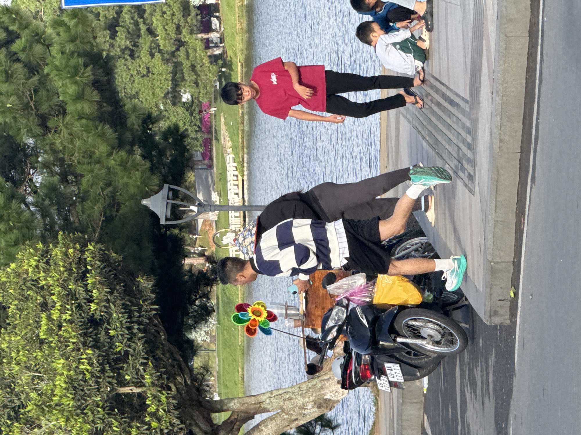 This image captures a street scene by Xuan Huong Lake in Da Lat, Vietnam. In the foreground, a black motorcycle is parked next to a wooden cart, which displays a colorful pinwheel. A man in a blue and white striped shirt and black shorts is walking away from the cart, holding a light blue object in his right hand. To his left, another person in dark clothing is partially visible. Further to the right, a young boy in a red t-shirt and black pants stands near the edge of the pavement, looking down. In the background, other children are crouched down, and a body of water, presumably Xuan Huong Lake, is visible, with trees and greenery lining its banks. The overall lighting suggests it is daytime, likely late afternoon given the shadows.