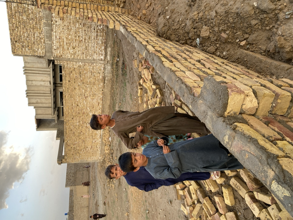 The image captures an outdoor construction scene in Kandahar, Afghanistan, under a partly cloudy sky, likely during late afternoon or early evening. In the foreground, a partially constructed wall of light-yellow bricks, cemented with dark grey mortar, runs diagonally from the lower right towards the center-left. To the immediate left of this wall, a scattered pile of loose, unmortared bricks rests on the uneven dirt ground.
Four children are positioned near the bricks and the nascent wall. From left to right: a young boy in a dark blue shalwar kameez and light sandals stands with his head turned right. Next to him, a younger boy in a grey shalwar kameez stands looking downwards, holding a small, indistinct object in his hands. Behind and slightly to the right of him, a young girl, mostly obscured, is visible, wearing a light-colored, possibly patterned garment. Further right, a teenage boy in a brown shalwar kameez with loose-fitting trousers stands with his hands on his hips, looking towards the right side of the frame.
In the mid-ground, multiple structures with completed light-yellow brick walls enclose vacant plots of dirt land. In the background, a more developed concrete building with a distinct ribbed, vertical facade and windows is visible. The ground throughout the scene is unpaved, consisting of dry, disturbed earth. Several additional, more distant figures in traditional attire can be seen walking or standing in the background.
