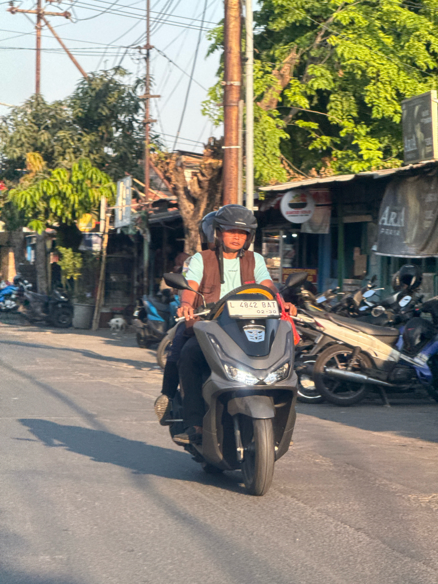 The image shows a man riding a dark gray scooter in a city street, likely in Surabaya, Indonesia, given the presence of the license plate format "L 4842 BAT 02-30," where "L" typically designates East Java plates. The man wears a helmet, a light blue shirt, and a brown vest. He is positioned in the foreground, facing the camera. Behind him, several other motorcycles are parked along the side of the street, indicating a busy urban environment. The street is paved, and the scene is illuminated by natural sunlight, casting shadows. Buildings, trees, and utility poles are visible in the background, consistent with a street in a residential or commercial area. The overall atmosphere is that of a typical day on an Indonesian street.