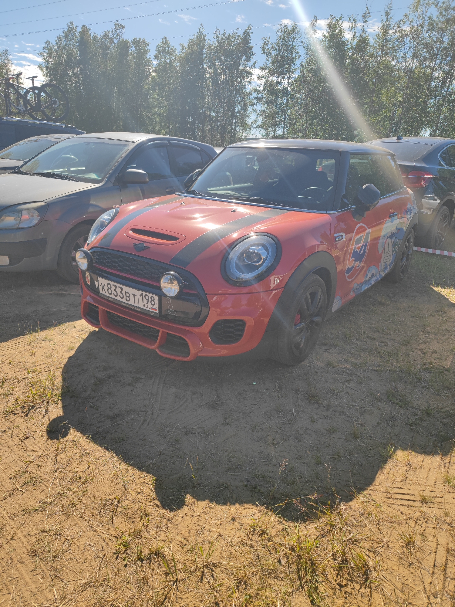A red Mini Cooper is parked on a sandy area with sparse dry grass. The car has black stripes on the hood, a custom decal on the side featuring the number "54" and a wave motif, and auxiliary lights mounted on the front grille. The license plate is visible, reading "K833BT 198 RUSSIA". A gray sedan is parked to the left of the Mini Cooper, and a black SUV is partially visible to the right. In the background, a line of trees stands against a bright blue sky with scattered clouds. The sun is shining brightly, casting shadows on the ground. The location is identified as Sertolovo, Russia.