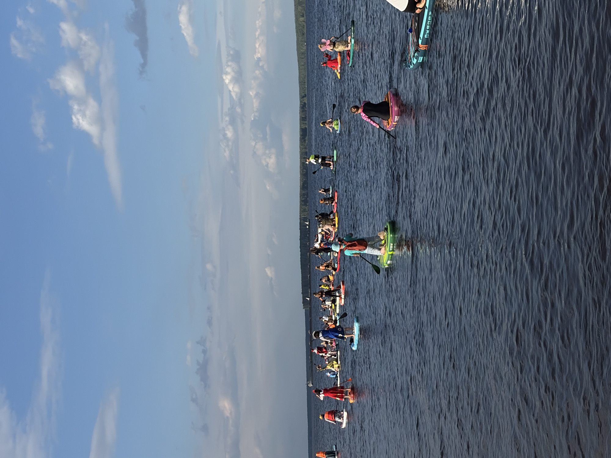 This media file depicts a large group of people stand-up paddleboarding on a calm body of water, likely a lake or river, near a tree-lined shore. The scene appears to be a recreational event or a gathering, with many participants spread across the water. The time of day seems to be late afternoon or early evening, as indicated by the lighting and the color of the sky, which is a clear blue with scattered white clouds. The water is dark and reflects the sky and surrounding environment.
The subjects are primarily individuals on paddleboards, dressed in casual attire suitable for water activities. Some are wearing life vests. A few individuals are dressed in costumes, including one person in a red Santa suit and another with fairy wings. The people are in various stages of paddling, with some actively using their paddles and others resting. The objects are the paddleboards themselves, which are diverse in color and design, and paddles.
Notable details include the sheer number of paddleboarders, suggesting a coordinated event. The backdrop of a distant, dense green forest hints at a natural, possibly remote, location. The water's surface shows gentle ripples, indicating a peaceful and serene environment. There is no discernible text visible in the image. The overall mood appears to be one of enjoyment and community, with people engaging in a shared outdoor activity. The setting is clearly Chaikovsky, Russia, as indicated by the provided location context.