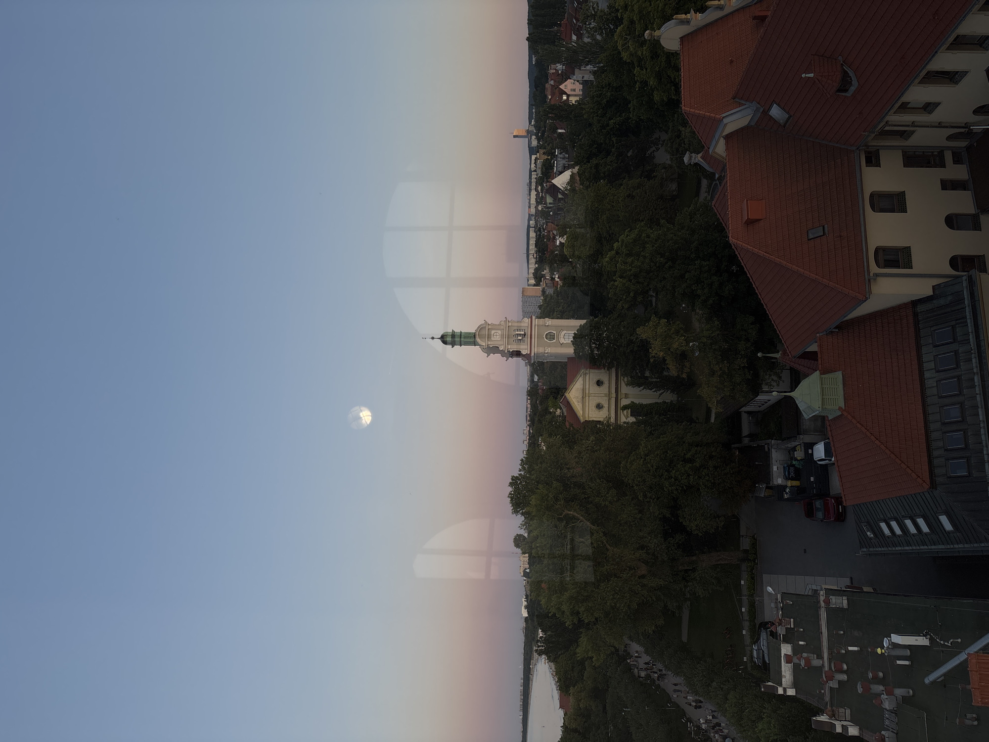 The image is an aerial view of the city of Sopot, Poland, taken at dusk. The sky is clear with a soft gradient of pink and blue. The moon is visible in the upper left quadrant of the sky. The foreground features several red-tiled roofs of buildings, with some windows visible. In the middle ground, there are lush green trees surrounding a building with a light-colored facade and a prominent steeple topped with a green dome. Further in the background, more buildings and trees are visible, extending towards the coastline. On the left side of the image, a body of water, likely the Baltic Sea, is visible, with a beach and a promenade lined with people walking. The overall scene depicts a peaceful evening in a coastal city.