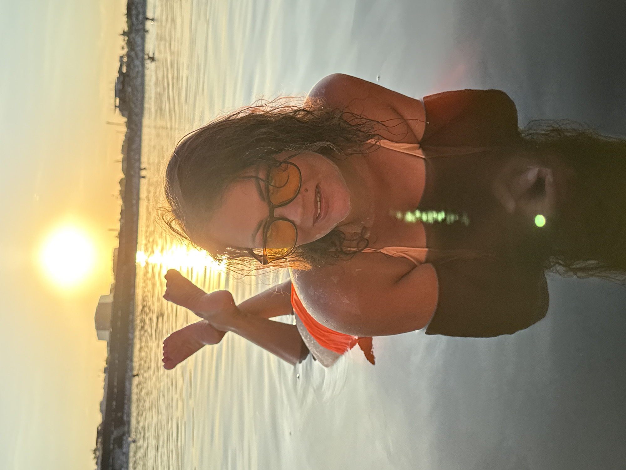 A woman is lying face down in calm water, her legs crossed and extended out of the water. She is wearing an orange bikini bottom and sunglasses. The sun is setting in the background, casting a warm glow over the scene. The woman appears to be enjoying the water. The location is the salt lakes of Sol-Iletsk, Russia, known for their therapeutic properties.