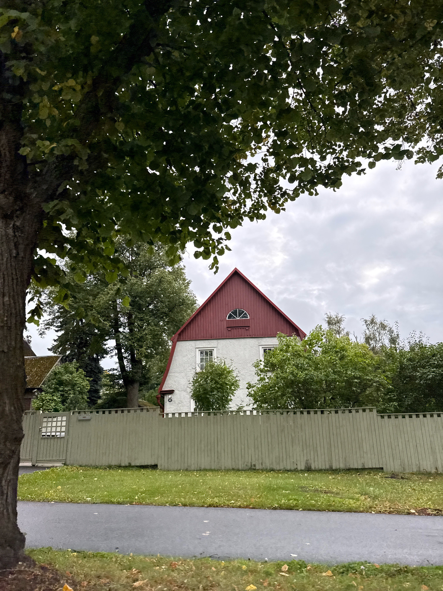 The image shows a residential property in Tallinn, Estonia, with a light-colored house featuring a red gable roof. The house has a small arched window in the gable and several standard windows. A light green wooden fence runs along the front of the property, with a gate visible on the left. A dark asphalt road borders the grassy area in front of the fence. Large trees with green leaves frame the scene from the foreground and background, indicating an outdoor, likely suburban or residential setting in Tallinn. The sky is overcast with clouds. There are no people visible in the image.