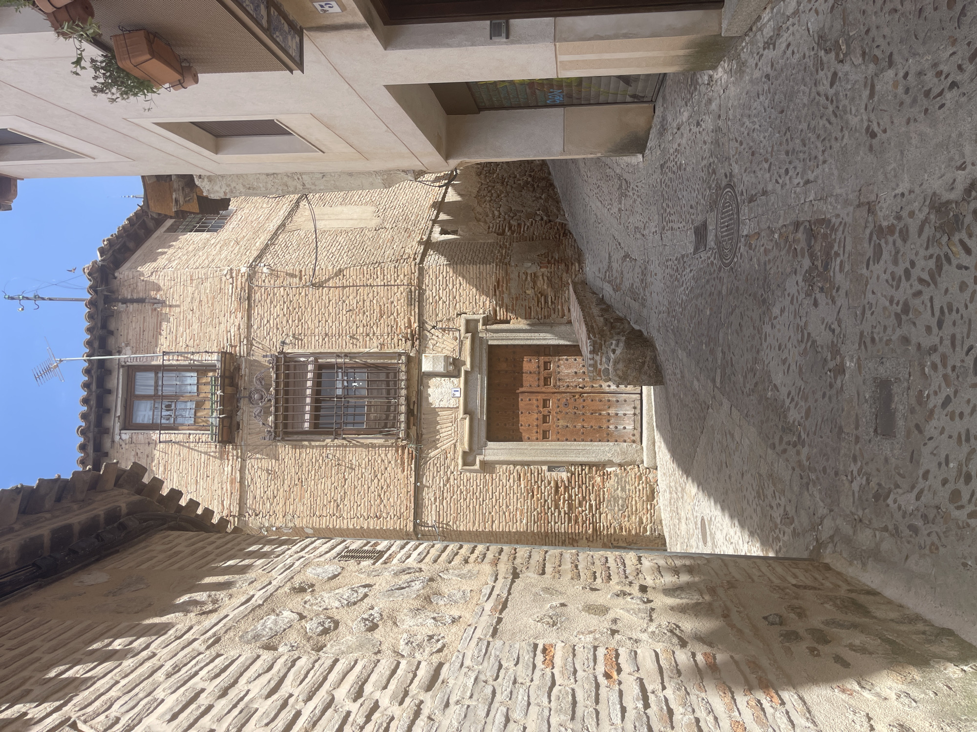The image depicts a narrow, cobblestone alleyway in the historic city of Toledo, Spain. The alleyway curves upwards and to the right, bordered by old stone and brick buildings. The building on the left is constructed with rough-hewn stones and rough plaster, with a tiled roof overhang casting a shadow. The building on the right is lighter in color, with a modern window and planters with greenery. In the center of the alley, a tall, multi-story brick building features a large wooden double door with studded metalwork and a smaller decorative archway above. The building also has barred windows and a small balcony. Sunlight streams down, creating strong shadows that define the textures of the cobblestones and brickwork. The sky is clear and blue. There are no people or vehicles visible in the scene.