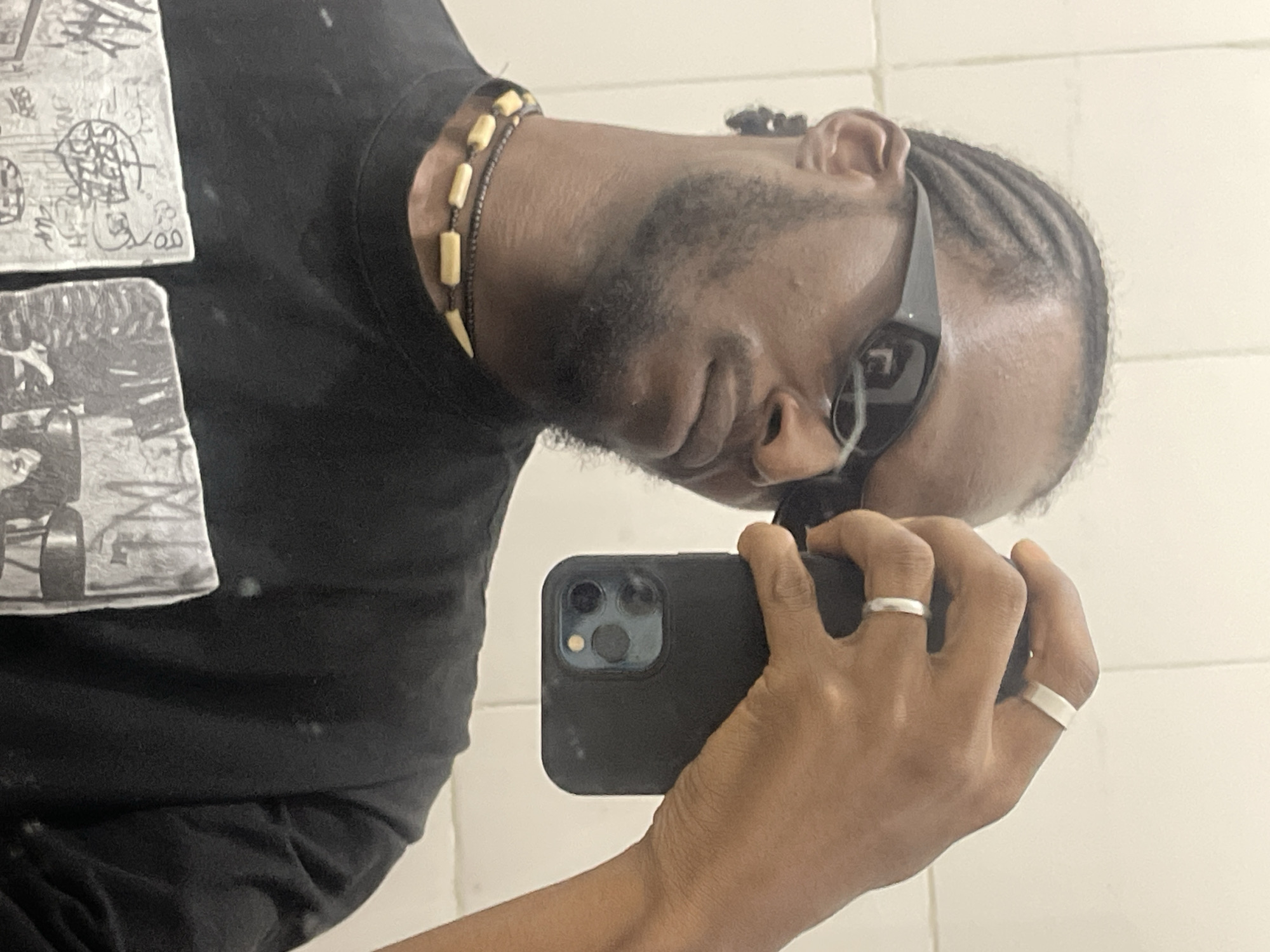 The image depicts a mirror selfie of a single individual, identified as male, of African descent, with dark skin, a beard, and cornrow braids. He is wearing dark, rectangular-framed sunglasses and a black t-shirt with a monochrome graphic print on the chest. Around his neck, he wears two layered necklaces: an upper one consisting of a dark cord strung with light-colored, elongated tubular beads, and a lower, thinner dark cord. His right hand is prominently featured, holding a dark-cased smartphone (identifiable by its triple camera array and flash) as if capturing his reflection. Two silver-toned rings are visible on his right hand: a wide band on the index finger and a thinner band on the ring finger. The background consists of a plain, light-colored wall, possibly tiled, indicating an indoor setting. The scene is stated to be from the city of Jos, Nigeria, though the visual content does not provide specific identifiers for this location.