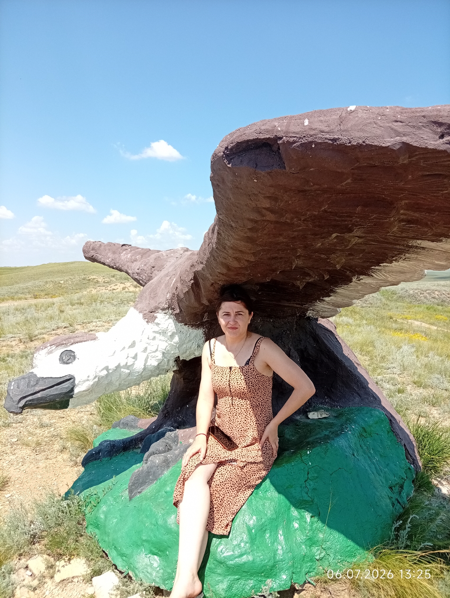 A woman is sitting on a large, colorful rock sculpture of an eagle in a grassy, semi-arid landscape under a blue sky with scattered clouds. The eagle sculpture is brown and white with a black beak and eye. The woman is wearing a leopard-print dress and is positioned on the green part of the rock. The background shows rolling hills and sparse vegetation. The timestamp "06.07.2026 13:25" is visible in the lower right corner of the image. This scene is located in the city of Aleksandrovka, Kazakhstan.