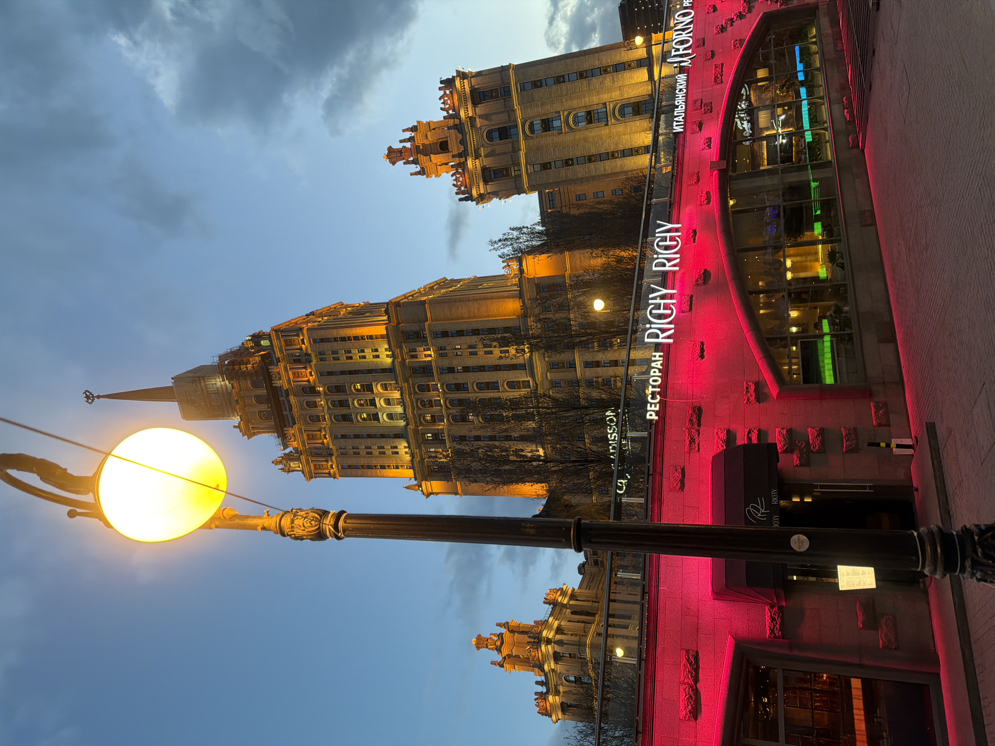 The image shows a nighttime view in Moscow, Russia, featuring Stalinist architecture and modern establishments. In the foreground, a vintage-style street lamp with a glowing spherical light is visible. In the middle ground, two impressive, illuminated Stalinist buildings dominate the skyline. Adjacent to one of the buildings is a modern restaurant illuminated with pink lights. The restaurant's name, "RICY RICHY," is prominently displayed in neon letters. The sky above is a mix of dark clouds and lighter blue hues, indicating a twilight or early evening setting. No people are clearly visible.