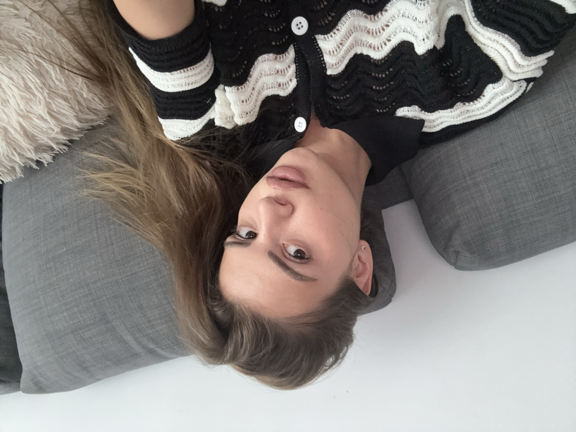 An overhead portrait features a young adult female lying down, looking directly at the camera. Her head is positioned towards the bottom of the frame. She has long, light brown hair flowing around her head. Her expression is neutral, with a direct and steady gaze. She wears light makeup, highlighting her lips and eyebrows, and a small stud earring is visible in her left ear.
She is dressed in a black and white cardigan or sweater characterized by a wavy, open-knit pattern, where the white sections exhibit a lace-like texture. The cardigan has white buttons down the front, and a plain black collar is visible beneath it.
The subject is resting on or surrounded by grey fabric cushions or pillows, suggesting she is on a sofa or bed. A fluffy, cream-colored pillow or blanket is partially visible near her hair on the left side. The lower background is a plain white surface, likely a floor or wall, indicating an indoor setting. There is no visible text, and the time of day or weather cannot be determined from the image.
