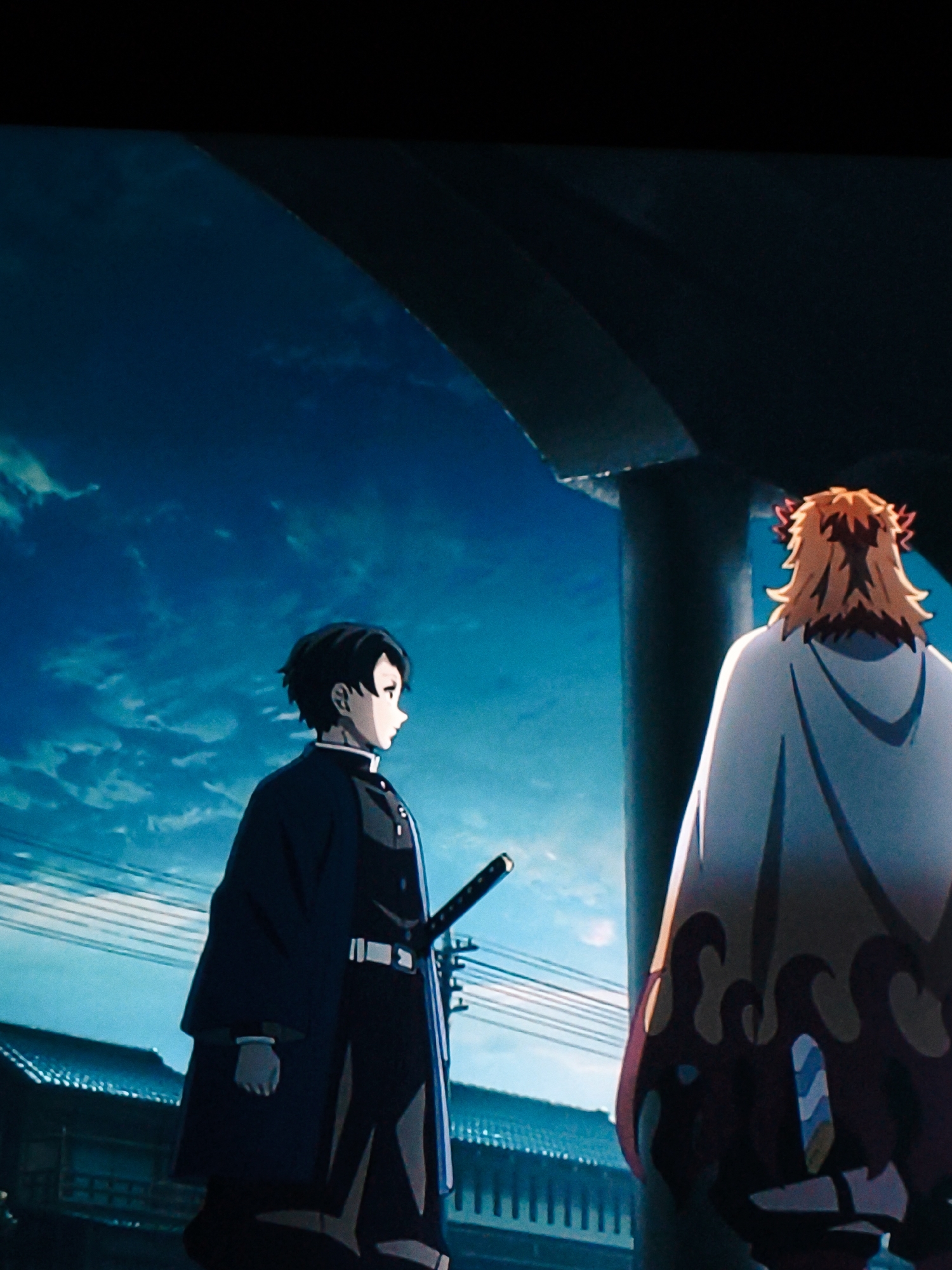 Two animated characters stand on a street, facing away from the camera. The character on the left has short black hair and wears a dark uniform with a white collar and belt, and a sword at his hip. The character on the right has long, spiky orange hair with red accents and wears a white hooded cloak with a flame-like pattern. The background shows a dark blue sky with scattered clouds, power lines, and traditional Japanese-style buildings. The scene appears to be set at dusk or night, with a large dark structure overhead, possibly part of a bridge or overhang. There is no discernible connection to the city of Vladivostok, Russia, as the architectural and stylistic elements suggest a setting in Japan.