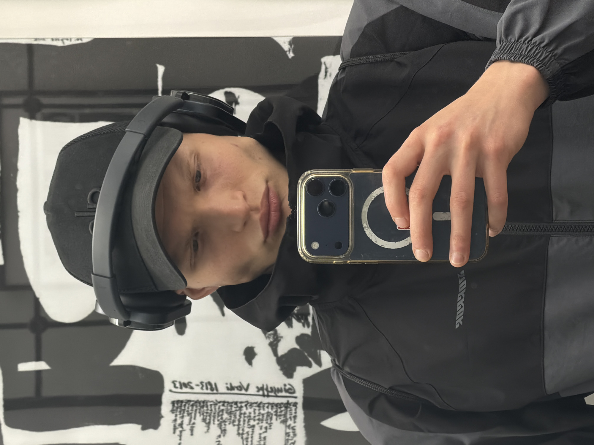 A person is taking a selfie with a smartphone. They are wearing a black hooded jacket with grey accents, a black baseball cap, and grey headphones. The person's hand is holding the phone, which has a clear case with a magnetic ring on the back. The background is a wall with black and white graphics, some of which appear to be text. The environment and style of the graphics suggest an urban setting, though without any additional landmarks it is not possible to confirm a specific location in London.
