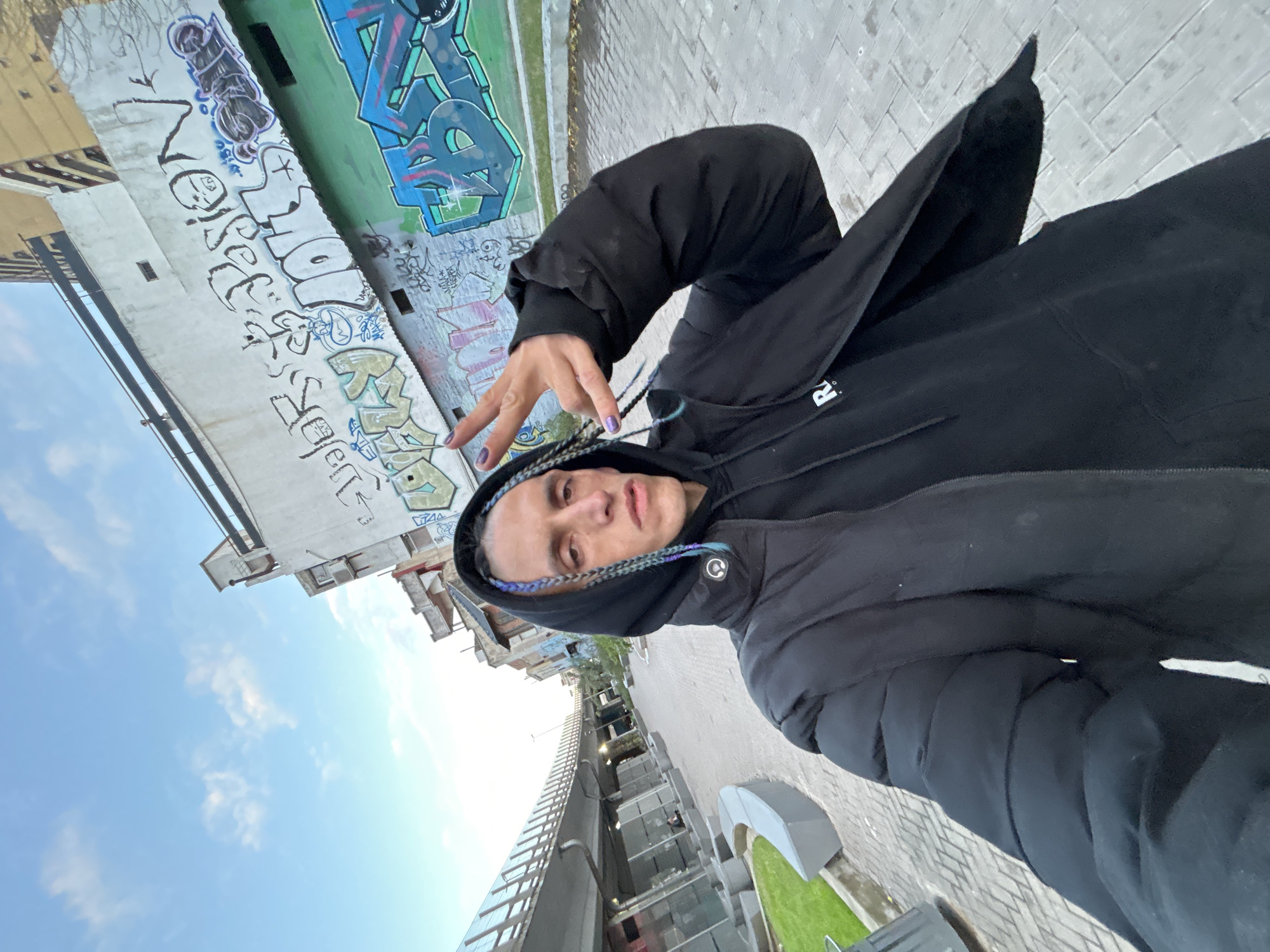 A person is taking a selfie in what appears to be Buenos Aires, Argentina. The person is wearing a black hooded jacket and has braided hair with colored strands. They are making a hand gesture, possibly a peace sign or similar.
The background includes buildings with graffiti, a partially cloudy sky, and what looks like a pedestrian overpass or walkway. The setting appears to be an urban environment with elements of modern construction combined with older, decaying buildings. The street is paved with brick.