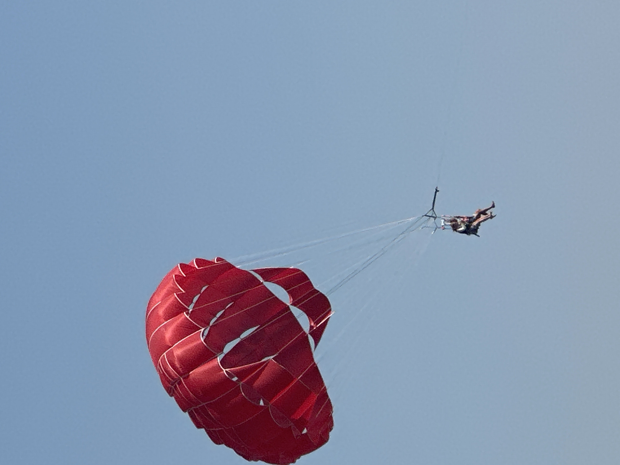 A bright red parasail, inflated and taut, is suspended in a clear, pale blue sky. Beneath it, two individuals, likely a tandem pair, are seated in a harness, appearing relaxed as they are pulled through the air. The parasail occupies the left side of the frame, its rounded shape and segmented design clearly visible. Faint white lines, possibly stitching or structural elements, are apparent on the surface of the red fabric. Thin, white lines, representing the tow ropes, extend from the parasail to the harness where the individuals are positioned. The individuals are dressed in swimwear or casual attire. The overall scene is one of leisure and airborne activity against a serene sky, indicative of a tourist resort or recreational area, possibly along the coast of Boreti, Montenegro, where such activities are common.
