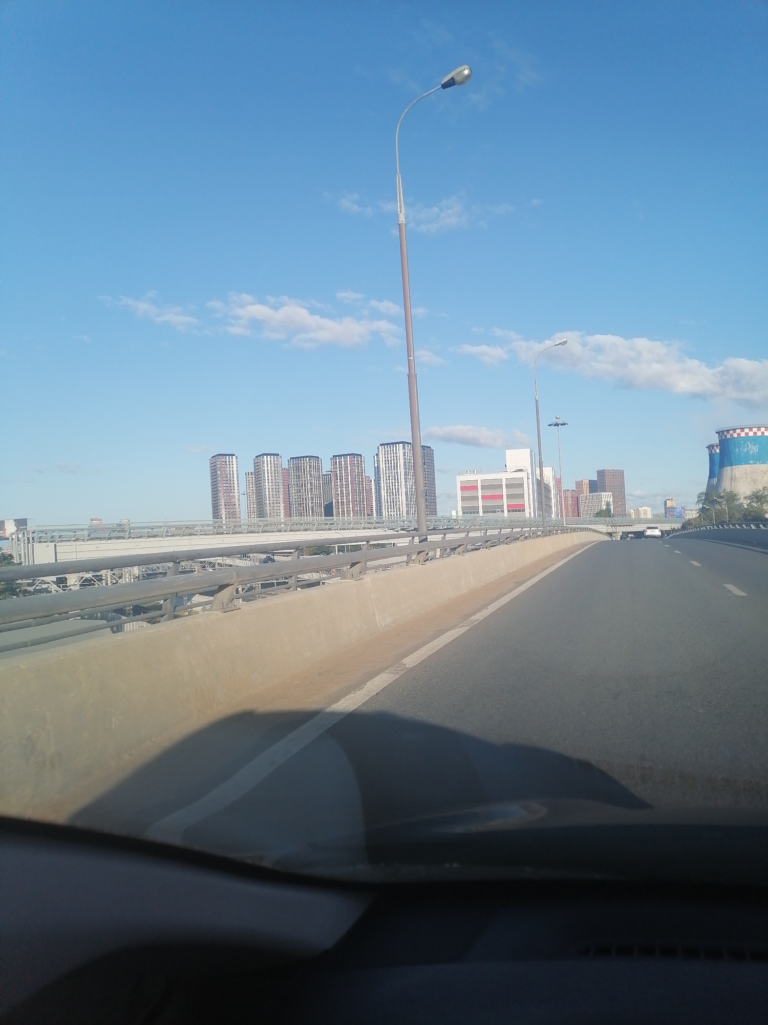 The image depicts a view from a vehicle on an elevated highway in Moscow, Russia. In the background, a cluster of modern skyscrapers with varied architectural designs, some featuring red accents, rises against a clear blue sky with sparse white clouds. To the right, a large industrial cooling tower with blue and red banding is visible. The highway itself is paved and marked with white lines, bordered by a concrete barrier. Streetlights are present on poles along the highway. The foreground shows the interior of a vehicle, with a dark dashboard and a shadow cast on the road.