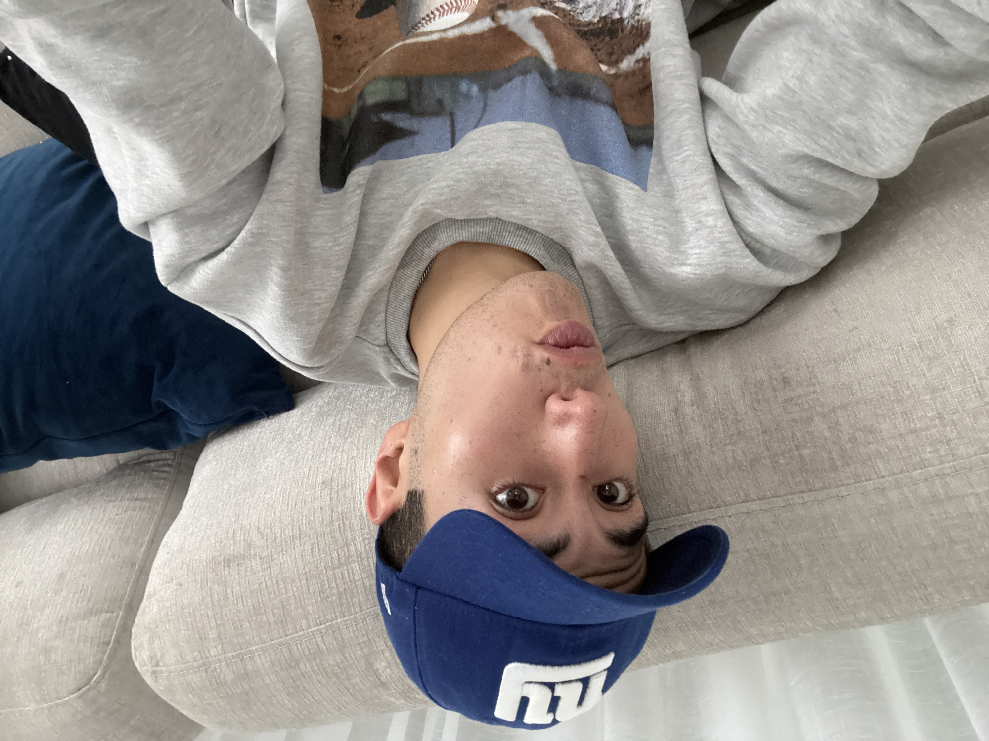 In an indoor setting, a young adult with dark hair and brown eyes is captured in a close-up, selfie-style photograph. They are wearing a blue New York Giants baseball cap and a gray crewneck sweatshirt with a graphic print of a baseball field and a baseball. The person is seated on a beige sofa, with a dark blue pillow visible behind them on the right. The lighting appears to be natural, coming from the left. The background suggests a domestic environment, with textured upholstery of the sofa and a pale curtain or wall on the far left. The specific location in the city of San Bernardino Tlaxcalancingo, Mexico, is not discernible from the visual information provided in the image.