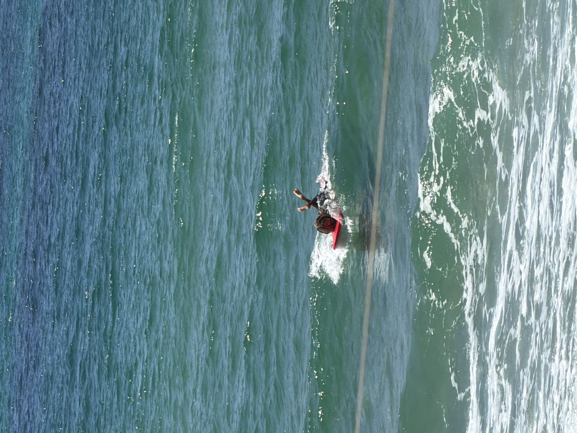 A person is bodyboarding in the ocean off the coast of Tel Aviv, Israel. The water is a deep blue-green and choppy with some white foam. The person is lying on a red bodyboard and appears to be moving through the water. The sun is shining, creating bright highlights on the water's surface. In the foreground, a wave is breaking, creating white foam that washes towards the shore.
