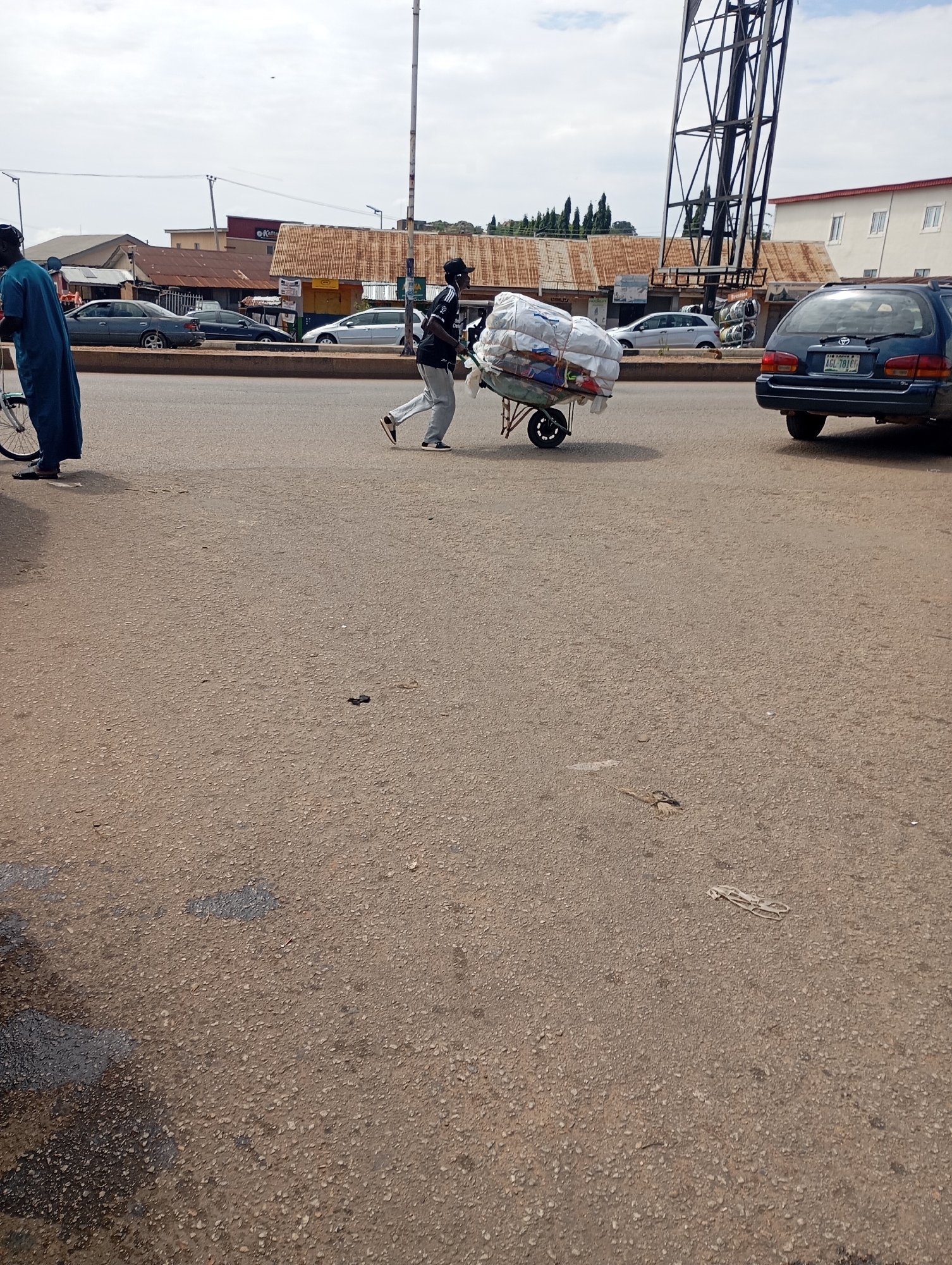 The image depicts a street scene in Jos, Nigeria, likely a commercial area with shops and ongoing traffic. In the foreground, the asphalt road is visible, with some debris and markings. To the left, a person dressed in a blue robe stands near a bicycle. Further to the right, a man is pushing a wheelbarrow laden with a large bundle of white materials, possibly clothing or textiles, wrapped in plastic and rope. He is wearing a black shirt with white stripes and a black cap. Behind him, several cars are visible, including a dark blue station wagon with a Nigerian license plate (AGL 701 CI) in the right lane. In the background, buildings with corrugated iron roofs and a white, multi-story structure can be seen. A tall metal tower, part of an electrical pylon, dominates the upper right portion of the image. The sky is overcast with some clouds. The scene suggests a bustling, everyday moment in the city.