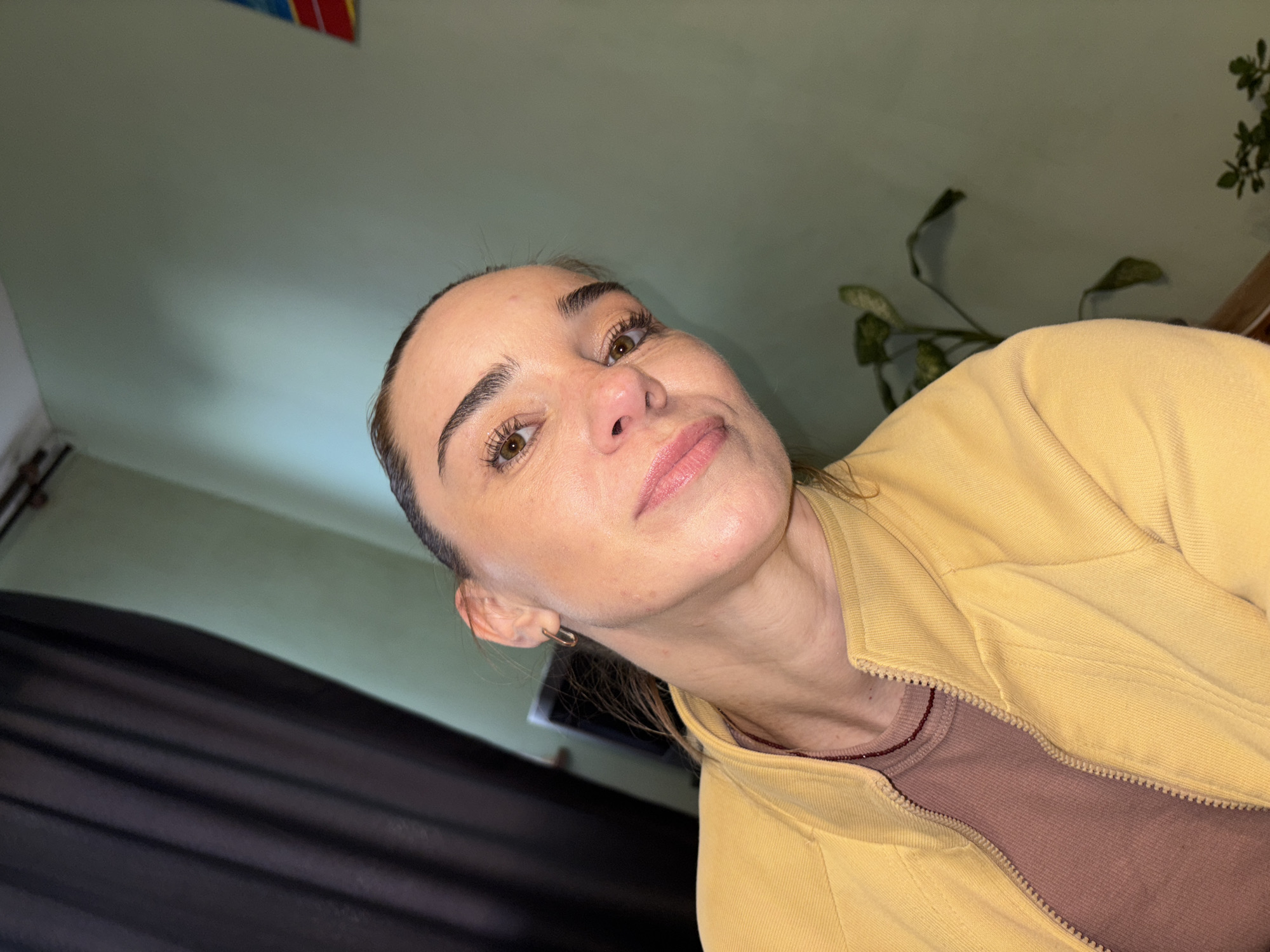 A woman with brown hair pulled back and wearing a yellow zip-up jacket over a brown t-shirt is captured in a close-up, slightly angled selfie. Her head is tilted to the side, and she is looking towards the camera with a soft smile. Her eyes are brown, and she is wearing subtle makeup. A thin gold hoop earring is visible in her left ear. The background features a dark curtain on the left and a pale green wall on the right, with a potted plant visible on the right side of the frame. There is no discernible indication of a specific city or country, or any further contextual details about the environment.