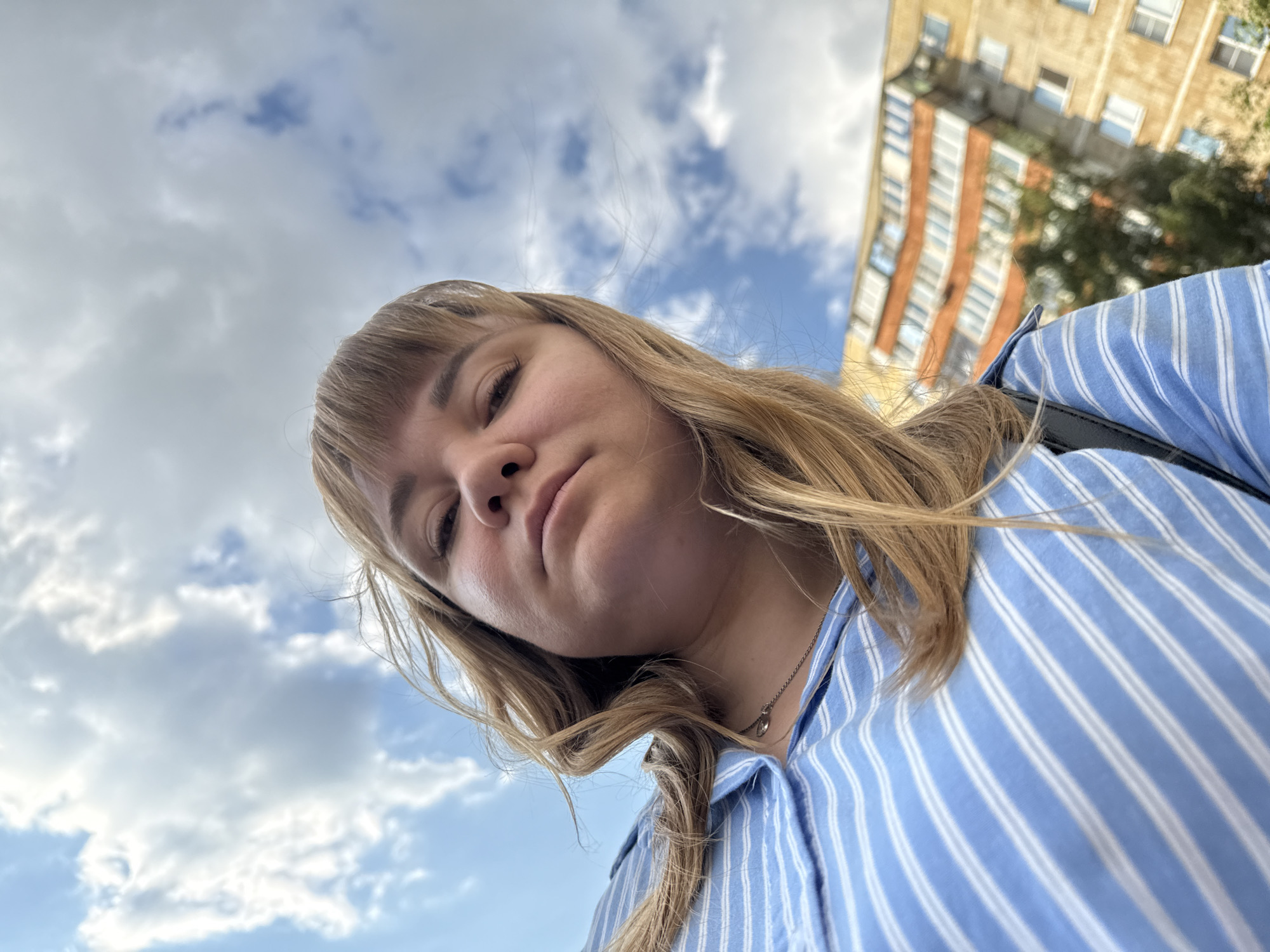 This media file is a low-angle, close-up selfie taken outdoors. The main subject is a young woman with blonde hair and bangs, wearing a blue and white striped button-down shirt and a thin silver necklace with a circular pendant. Her head is tilted downwards, and she is looking towards the camera with a neutral expression.
In the background, a cloudy sky with patches of blue is visible. To the right, a multi-story building with orange and beige brickwork and numerous windows can be seen, suggesting an urban or suburban setting. The lighting indicates it is daytime, likely late morning or afternoon, under partly cloudy conditions. The perspective of the selfie, looking up at the woman and the sky, gives a sense of looking up from a low vantage point.
The image does not contain any visible text. The overall mood is casual and candid, typical of a selfie taken spontaneously. The location context provided, Stary Oskol, Russia, places the scene in a specific geographical area.