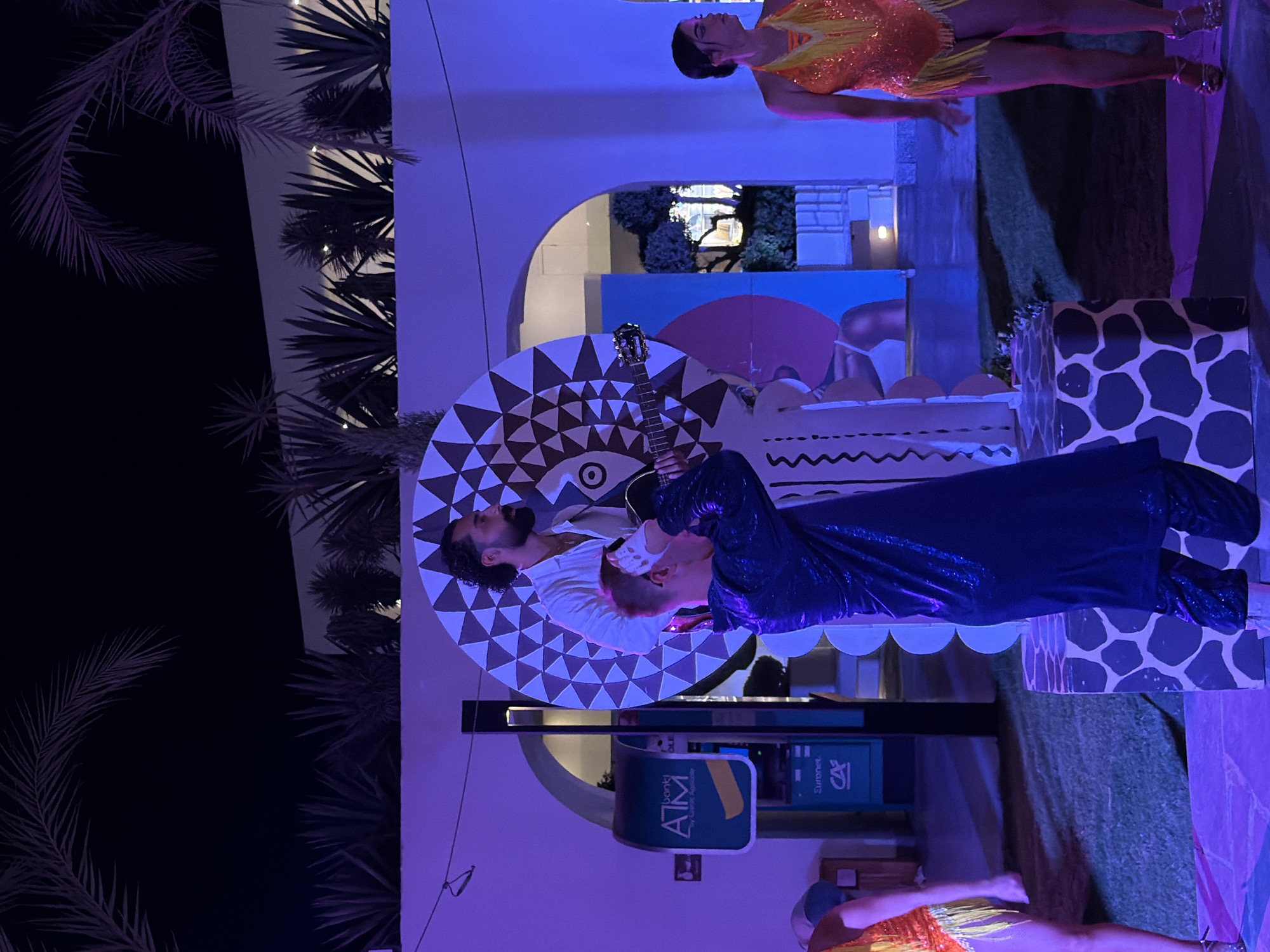 A man with dark, curly hair and a beard sings into a microphone while playing an acoustic guitar. He is wearing a white shirt. Beside him, another person with red hair, dressed in a sparkly blue outfit, leans in towards him. They are standing on a circular platform decorated with a geometric pattern. In the background, a woman in an orange, sequined dress with yellow fringe is suspended in the air, with her hand extended. The scene is illuminated by purple lighting, and palm trees are visible on the left side of the image. The overall setting appears to be an outdoor evening event. There is an ATM machine visible with text that includes "bank", "ATM", "by Crédit Agricole", and "CA". This is likely a performance or entertainment event in Sharm El Sheikh, Egypt, given the palm trees and warm-toned lighting.