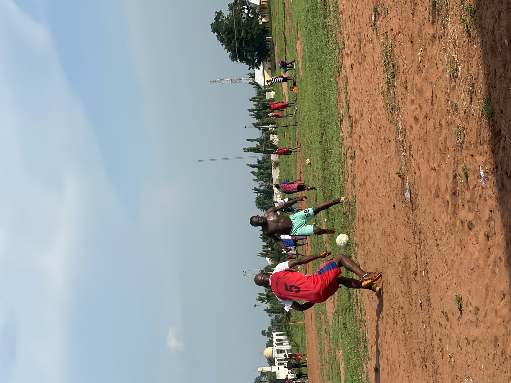 The image depicts an outdoor football (soccer) match or practice session taking place on a sunny day in Ogbogbo, Nigeria. The scene is dominated by a playing field composed of reddish-brown dirt and scattered patches of green grass.
In the foreground, two male players are actively engaged in play with a white football. One player, shirtless and appearing to be sweating, wears light green shorts with the number "89" visible on them. He is positioned on the left side of the frame. The second prominent player, on the right, is wearing a red and white jersey that clearly displays "OGBOGBO FC" and the number "5" on the back, paired with blue and red shorts. Both players are in active stances, focused on the ball.
Several other men are visible further back on the field, some standing and others seemingly participating in the game. The background features a line of trees, various buildings, and a distinct white building with a dome and minarets, characteristic of a mosque. A tall, red and white communications tower is also visible in the distance. The sky above is light blue with some faint clouds, indicating clear weather and a bright time of day, likely midday or early afternoon due to the strong illumination.