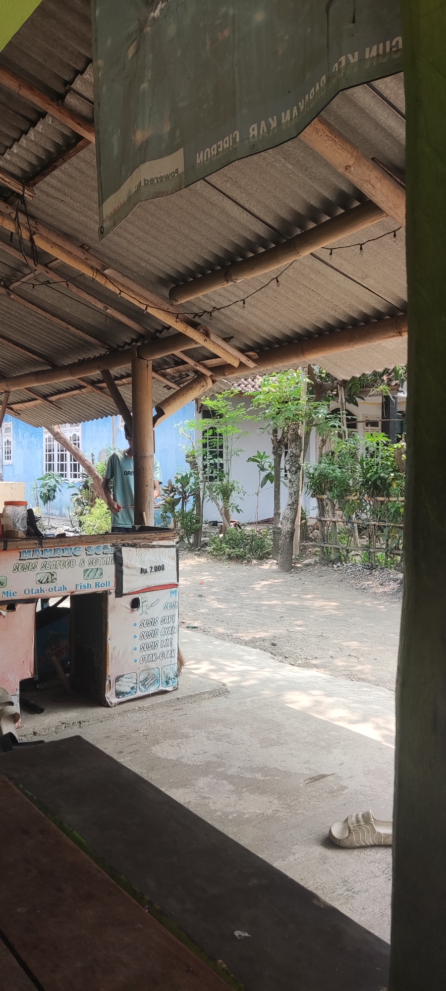 The image depicts an outdoor scene under a corrugated metal roof supported by bamboo poles. In the foreground, a wooden table is visible. To the left, a food stall with a pastel-colored front displays various menu items. One item listed is "Rp. 2.000". The stall also features images of food and text in Indonesian, including "SOSIS SEAFOOD & SOTANG", "Mie Otak-otak", "Fish Roll", "SOSIS SAPI", "SOSIS AYAM", "SOSIS ME", and "OTAK-OTAK". A person wearing a light blue t-shirt is standing behind the stall, appearing to be preparing food. In the background, a blue building with white-trimmed windows is visible, surrounded by lush green foliage. The ground is a dirt path. A single white sandal lies on the concrete ground to the right. A banner with Indonesian text, partially obscured and reversed, is hung from the roof. The scene is likely located in Karangwangun, Indonesia, given the presence of Indonesian language signage and the typical architecture for outdoor food stalls in the region.