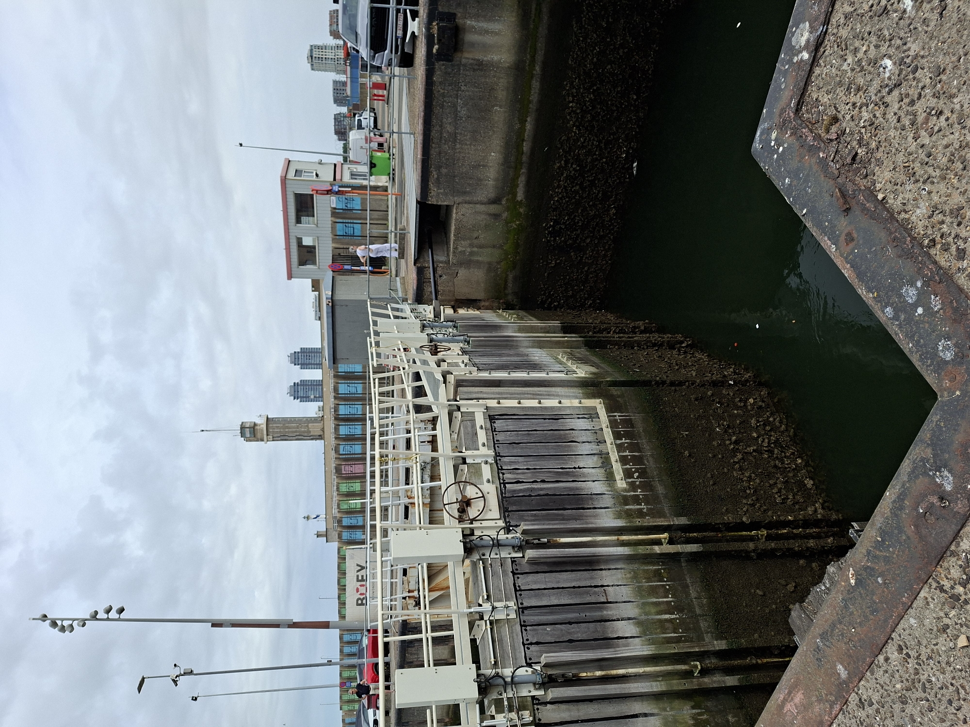 The image depicts a lock gate in the port of Ostend, Belgium. The lock mechanism is made of weathered wood and metal, with a large wheel for manual operation. The water inside the lock is dark green and murky, with visible growth on the lock walls. In the background, a white building with a red roof and two windows stands near a row of colorful, graffiti-covered concrete structures. A tall tower with a control room on top is visible further in the distance, along with several modern high-rise buildings. Two people are present: one man in a black shirt and shorts is walking away from the camera on the left side of the lock, and another person in white clothing stands on the walkway to the right. A red car is parked behind the man, and a silver SUV with a license plate is parked to the right. The sky is overcast, suggesting a cloudy day. The foreground shows a rusty metal edge of the lock structure.