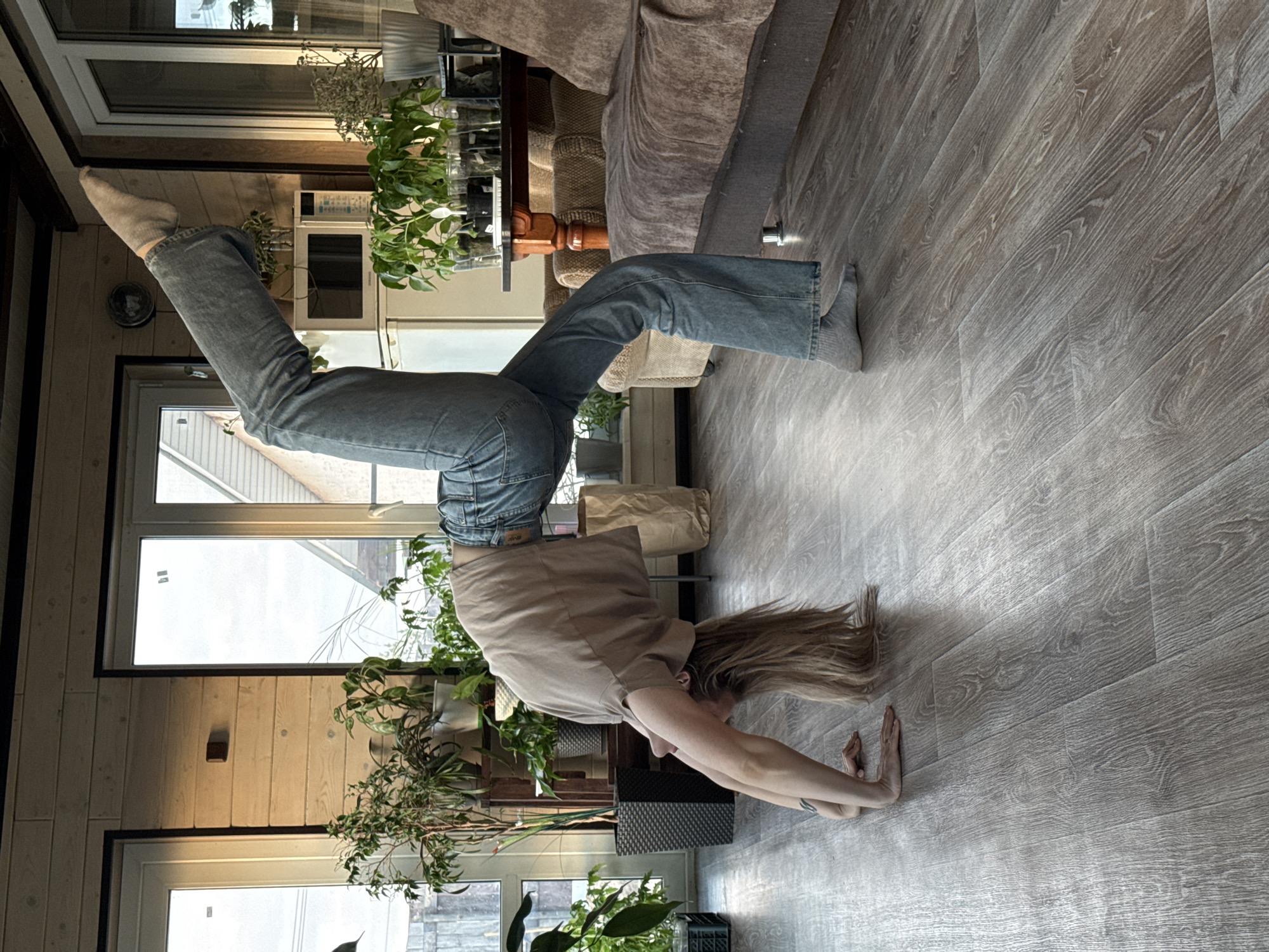 The image displays a person performing a handstand or headstand indoors, with their body inverted. The individual is positioned with their head near the floor and hands placed flat on the light-colored wooden laminate for support, while their legs are extended upwards, wearing light blue/gray denim jeans, a loose-fitting light brown short-sleeved top, and light gray socks. Long, light brown hair cascades towards the floor.
The setting is a well-lit room, likely a living space, characterized by ample natural light streaming through large windows in the background. The walls feature light-toned wooden paneling, and several potted plants are arranged throughout the space on shelves and tables, adding greenery. A dark brown sofa and a wooden table are also visible. The flooring exhibits a distinct wood grain pattern. The bright illumination indicates it is daytime. No discernible text is visible on any objects or clothing. The scene conveys a comfortable and naturally lit interior environment in Chaikovsky, Russia.
