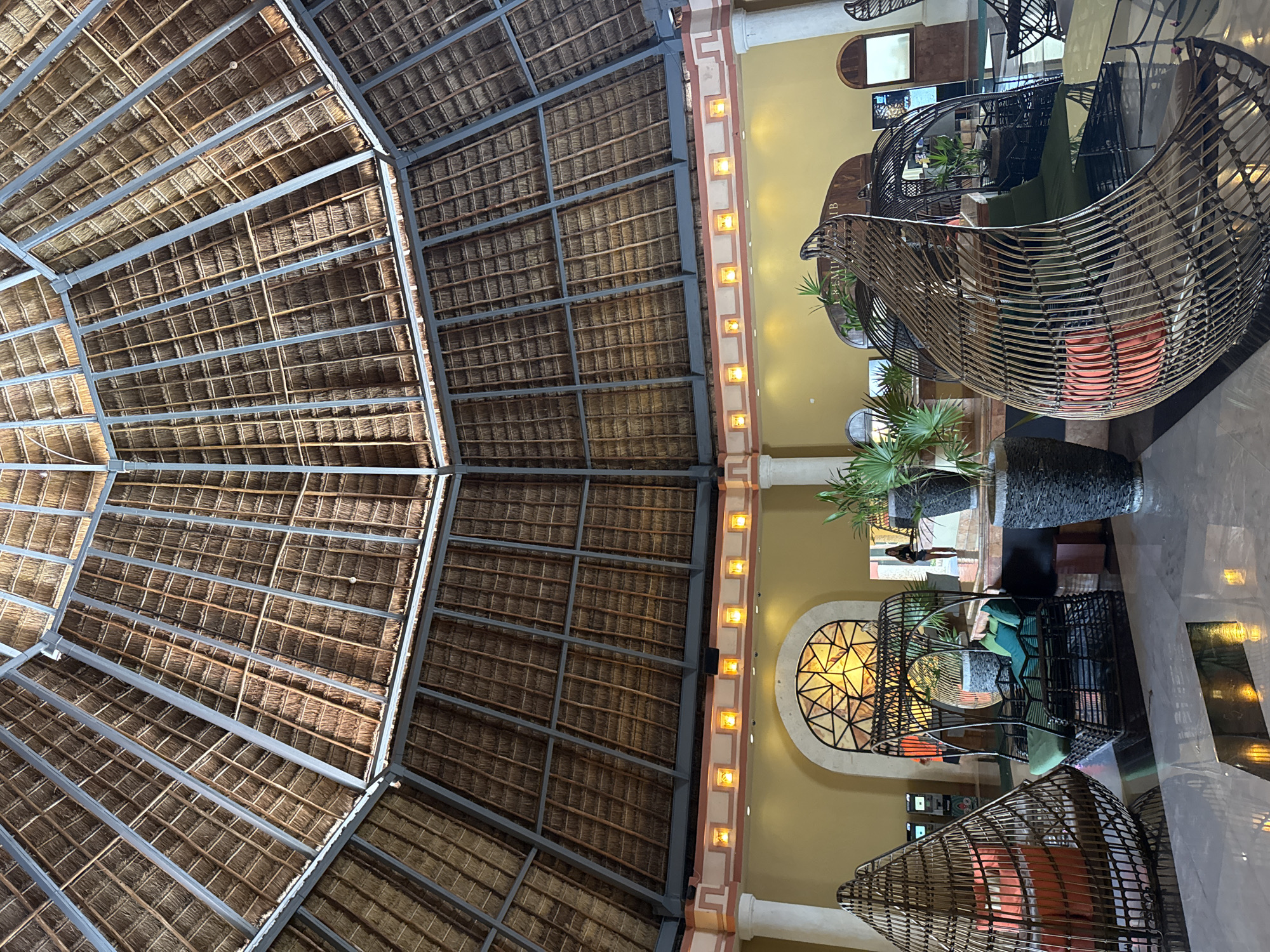 The image shows an indoor space, likely a resort lobby in Playa del Carmen, Mexico, given the architectural style. The most prominent feature is the thatched roof structure, supported by a grid of metal beams. The roof is divided into distinct sections, suggesting a large, multi-tiered construction. Below the roof, a pale yellow wall runs vertically, punctuated by a series of square sconces and decorative architectural details. Wicker or rattan-style hanging chairs are visible on the right side of the frame, with cushioned seats. Potted plants are positioned near the chairs. The overall atmosphere appears to be of a high-end tourist destination. The floor is marble or polished stone, reflecting light. No people are explicitly visible.