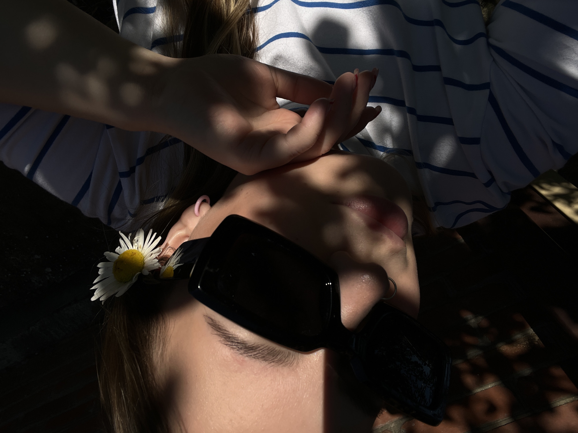 A close-up, high-angle, outdoor shot shows a person wearing black rectangular sunglasses and a white t-shirt with blue horizontal stripes. A white daisy with a yellow center is tucked behind the person's right ear. The person's left hand is resting on their face, obscuring their mouth and nose. The sunlight creates strong shadows across the person's face and body, as well as on a brick wall in the background. The scene is set in Novopokrovskaya, Russia.