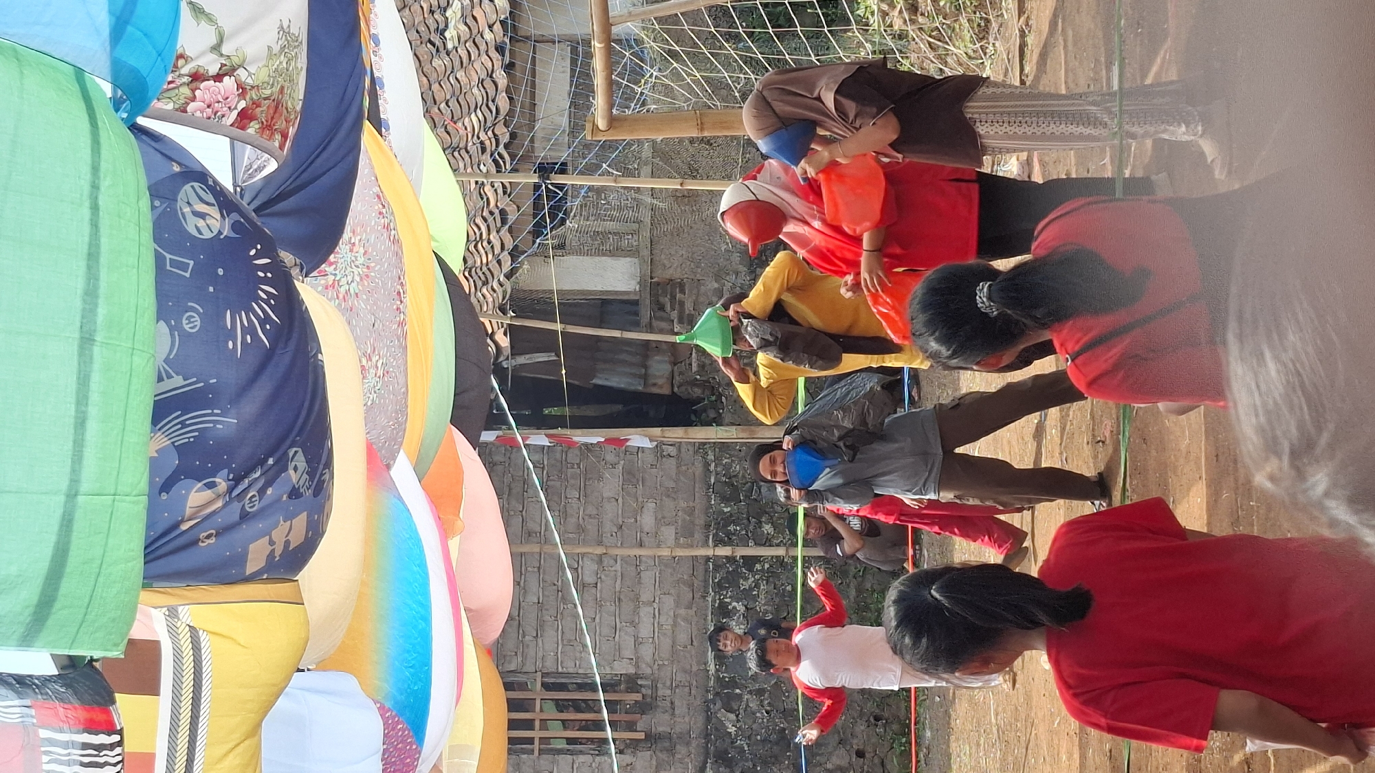 A group of people are participating in an outdoor activity under a canopy of colorful fabrics. The activity appears to be some kind of game or competition, as some individuals are wearing oversized, novelty items on their heads, such as a green cone-shaped hat and a red cone-shaped mask. One person is holding a black trash bag, and another is holding a blue funnel up to their face. The participants are dressed casually, and the setting seems to be a communal or recreational space. In the background, there is a brick building, a wooden fence, and a soccer goal. The ground is dirt. The colorful fabric canopy overhead suggests a festive or celebratory atmosphere. The overall scene suggests a community event taking place in a semi-urban or rural setting in Cilaku, Indonesia.