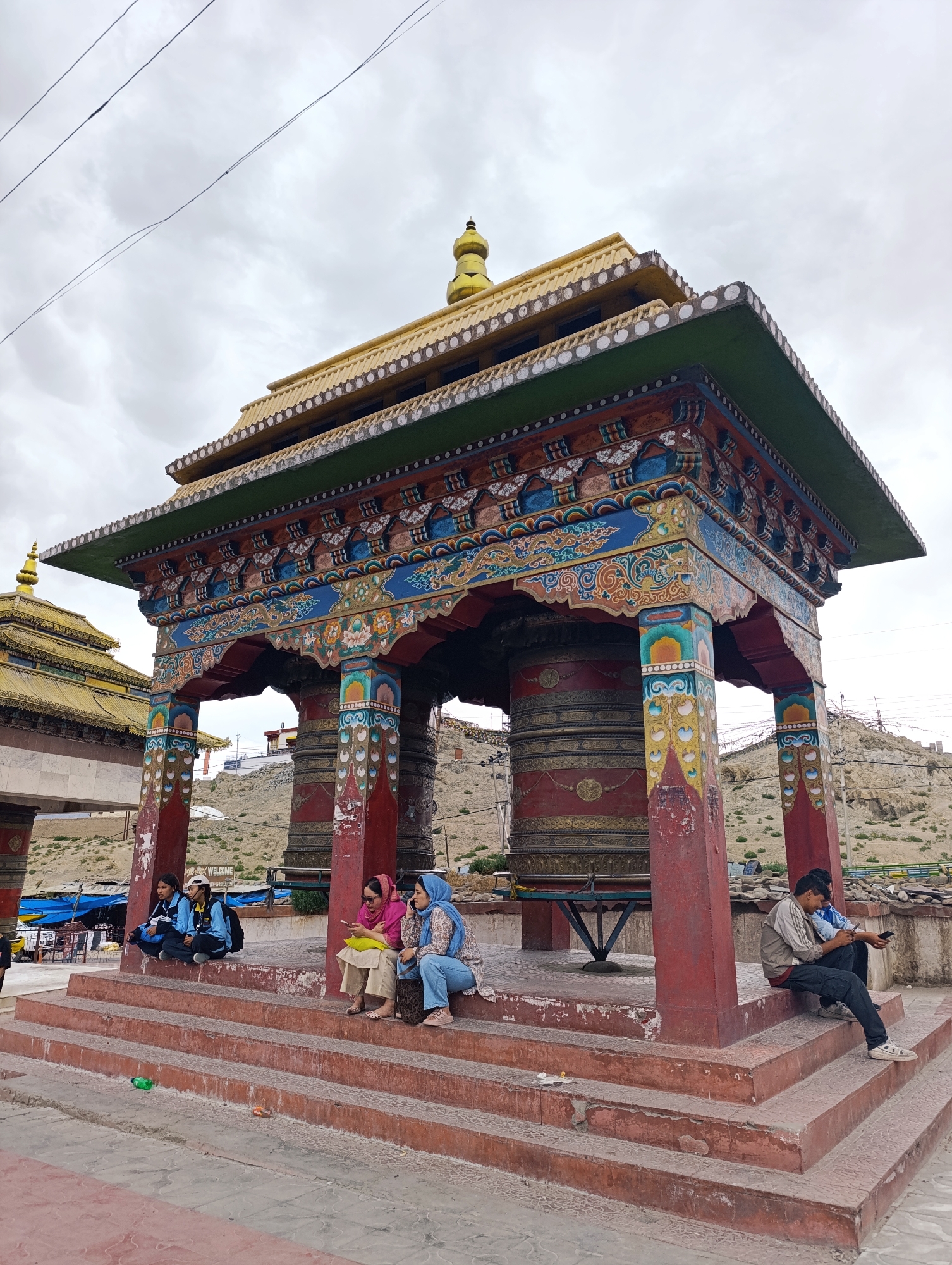 The image depicts a vibrantly painted Buddhist prayer wheel structure in Leh, India. The structure is ornate, featuring intricate carvings and colorful motifs on its pillars and roof. Two large, cylindrical prayer wheels are visible within the structure. In the foreground, several individuals are seated on the steps leading up to the structure. On the left, two young women are sitting together, one wearing a blue jacket and the other a yellow jacket and a baseball cap. To their right, two women are seated, one in a pink hijab and the other in a blue hijab. The woman in the pink hijab is looking at her phone, while the woman in the blue hijab is looking to the side. On the right side of the image, a young man wearing a light-colored jacket and dark pants is seated, looking at his phone. Behind the structure, a barren, hilly landscape is visible under a cloudy sky. The overall scene conveys a sense of cultural and religious significance.