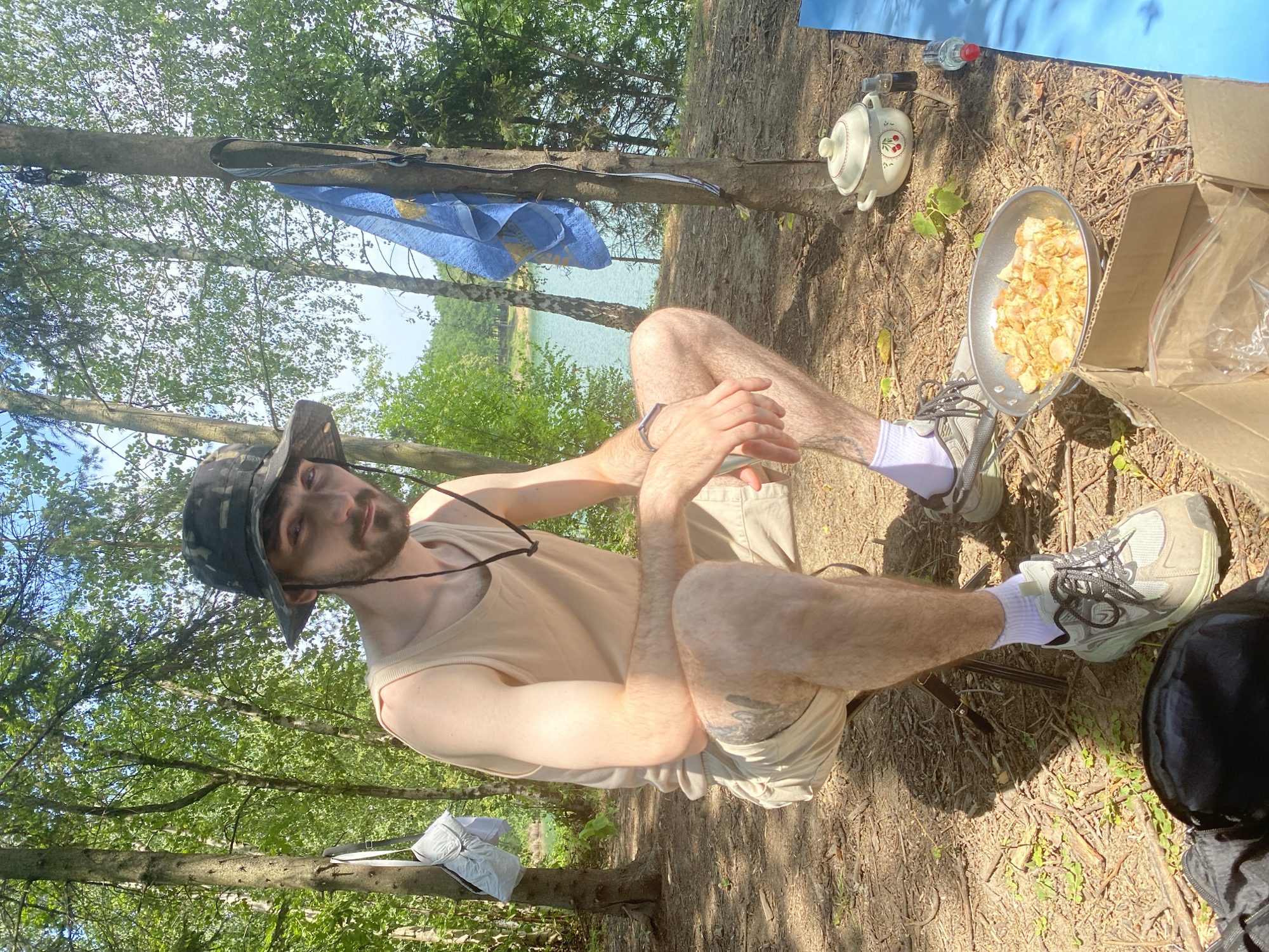 A young man wearing a camouflaged bucket hat, a light-colored tank top, and tan shorts is crouched on a folding chair outdoors. He has a beard and appears to be looking at the camera. In front of him, on the ground, is a frying pan filled with cooked food, possibly chicken. A cardboard box with plastic bags sits beside the pan. In the background, a body of water is visible, surrounded by trees. A blue towel hangs on a tree to the right of the man. The scene appears to be a campsite in or near Kaniów, Poland, given the context provided.
