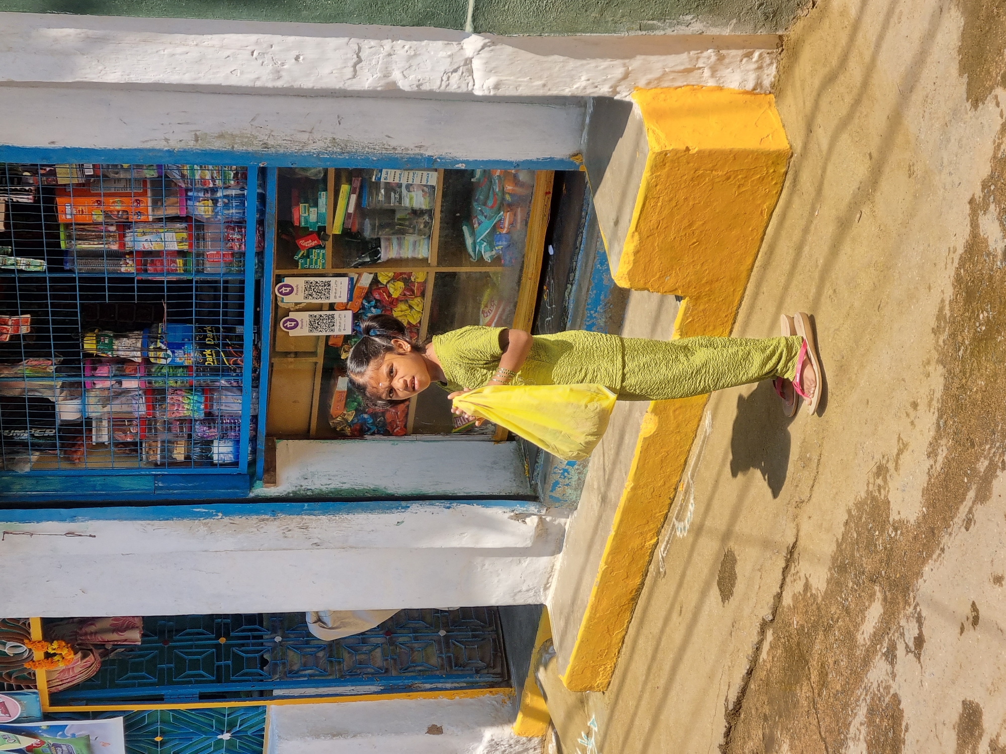 A young girl stands on a cracked concrete surface in front of a building in Pedda Kammavaripalli, India. She is positioned slightly right of center, facing the camera directly, wearing a textured, light green two-piece outfit and pink flip-flops. Her dark hair is styled in two pigtails, and a red mark is visible on her forehead. She holds a yellow plastic bag with both hands in front of her midsection.
Behind the girl, the building features a white painted facade with blue accents around openings. To her right, a shop window with blue metal grating displays an assortment of packaged goods, including food items and household products, on interior shelves. Two PhonePe QR code payment signs are clearly visible within the shop display. To her left, a blue metal gate with a geometric pattern is partially open, revealing a dark interior space. A marigold garland hangs above this gate. A yellow painted curb or raised platform runs along the base of the building, separating the storefront from the ground. Faint white chalk-like patterns are visible on the concrete near the curb. The ground shows patches of discoloration and distinct shadows from overhead objects and the girl herself, indicating bright sun exposure.