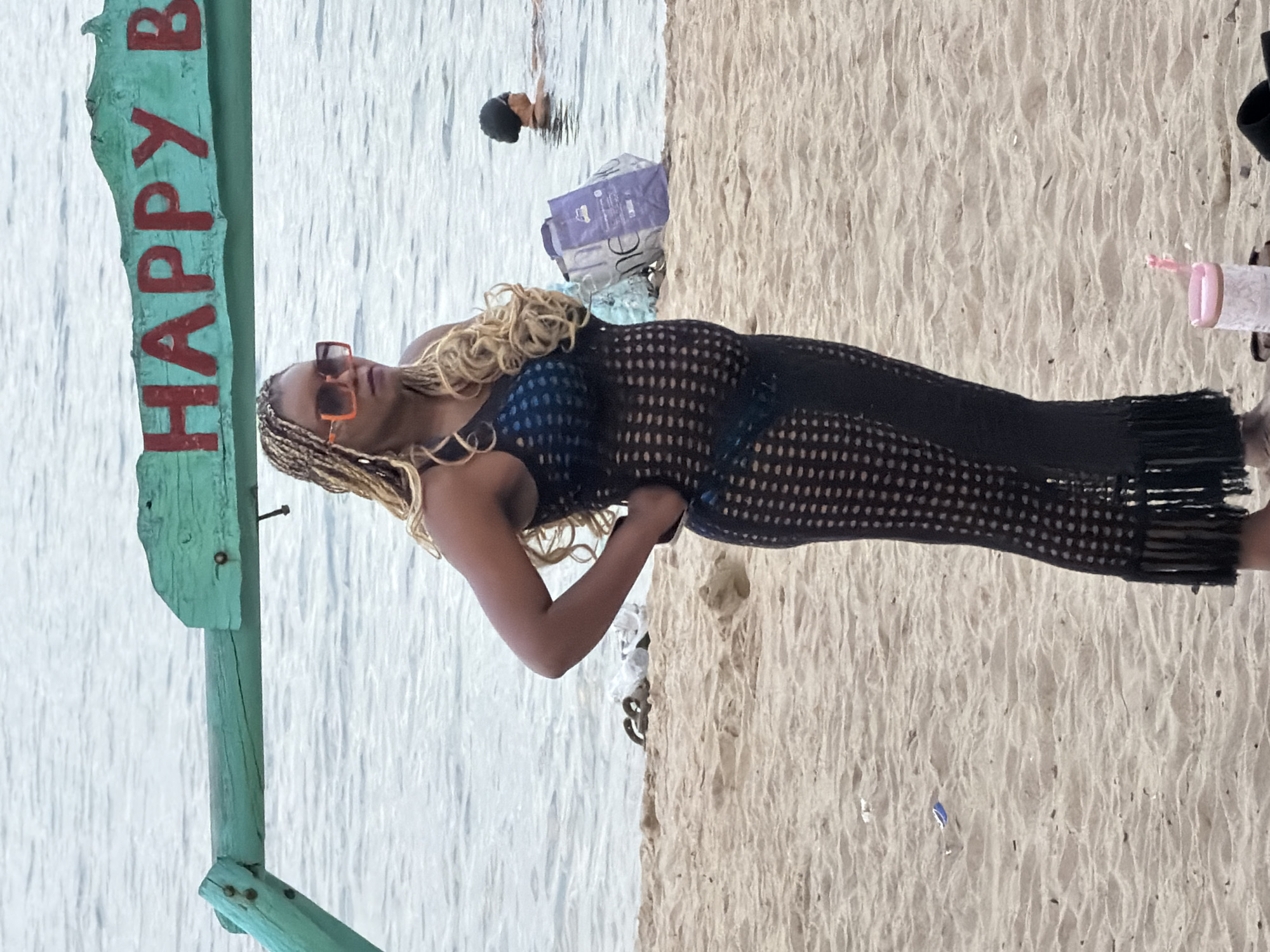 A woman stands on a sandy beach in Nha Trang, Vietnam, wearing a black crochet dress over a blue swimsuit and orange sunglasses. Behind her, the ocean stretches out, with a few people swimming in the distance. A wooden archway with "HAPPY B" painted on it in red letters is visible above the woman. The beach is littered with a few plastic items and a purple bag.
