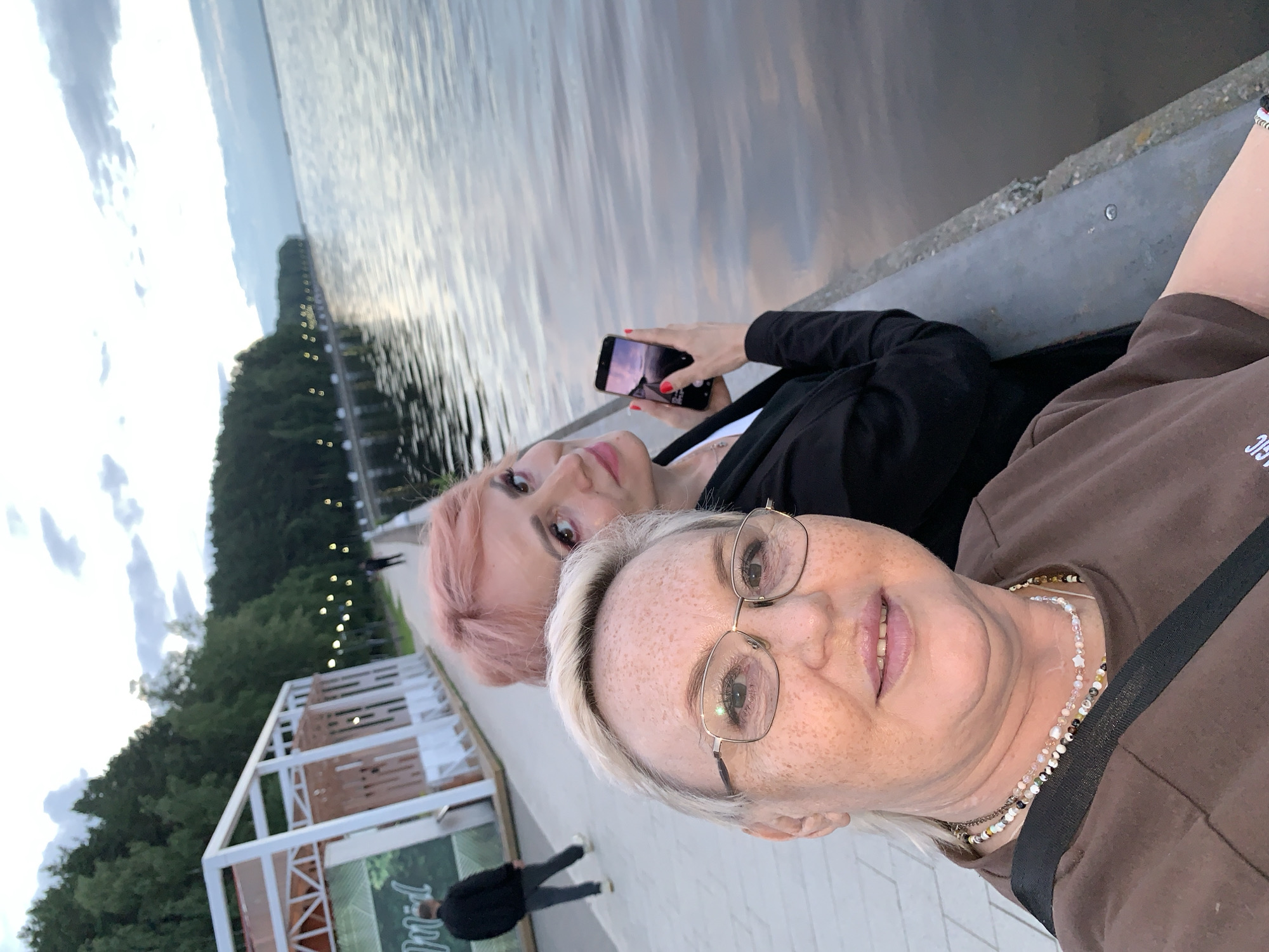Two women are captured in a selfie on a paved walkway next to a body of water, likely the Kama River, in Chaikovsky, Russia. The woman in the foreground, wearing glasses and a brown t-shirt with beaded necklaces, faces the camera directly and smiles. Behind her, another woman with pink hair, wearing a black jacket over a white shirt, holds up a smartphone to take the photo. Her eyes are directed towards the phone. In the background, a forested shoreline with a line of lights stretches along the river. A white gazebo-like structure with a red roof is visible to the left, and a person in a dark jacket walks away from the camera on the walkway. The sky is overcast with a mix of clouds and twilight hues.