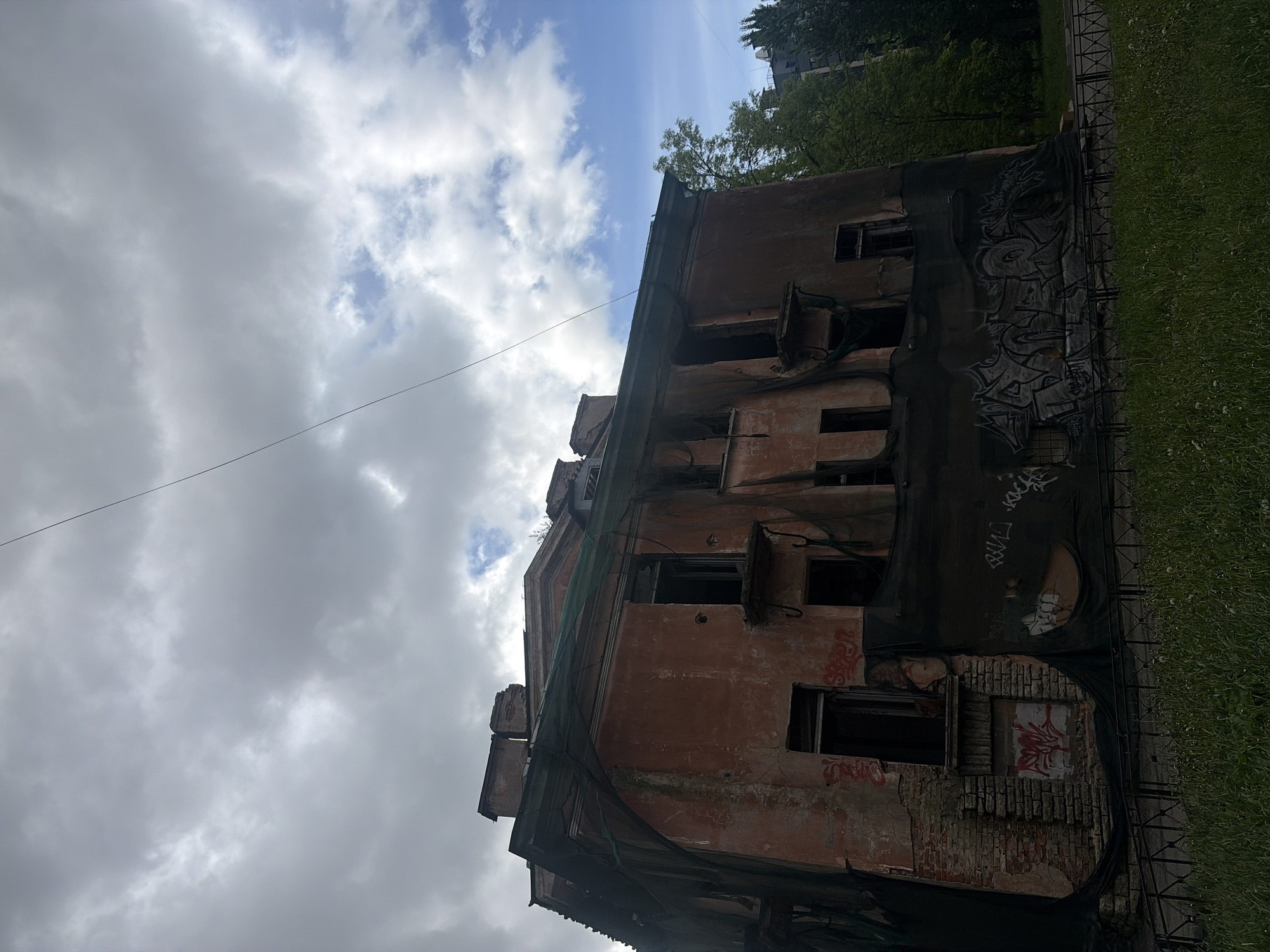 The image shows a decaying, multi-story building in Saint Petersburg, Russia. The building's facade is a faded reddish-brown, and it is partially covered with a dark mesh material bearing graffiti. Many windows are open and without glass. The building appears to be in a state of disrepair, with exposed brickwork. To the right of the building is a strip of green grass, indicating a park or lawn. The sky above is cloudy, with patches of blue visible. A powerline extends across the sky, partially obscuring the view. The building is oriented diagonally in the frame, capturing the full height.