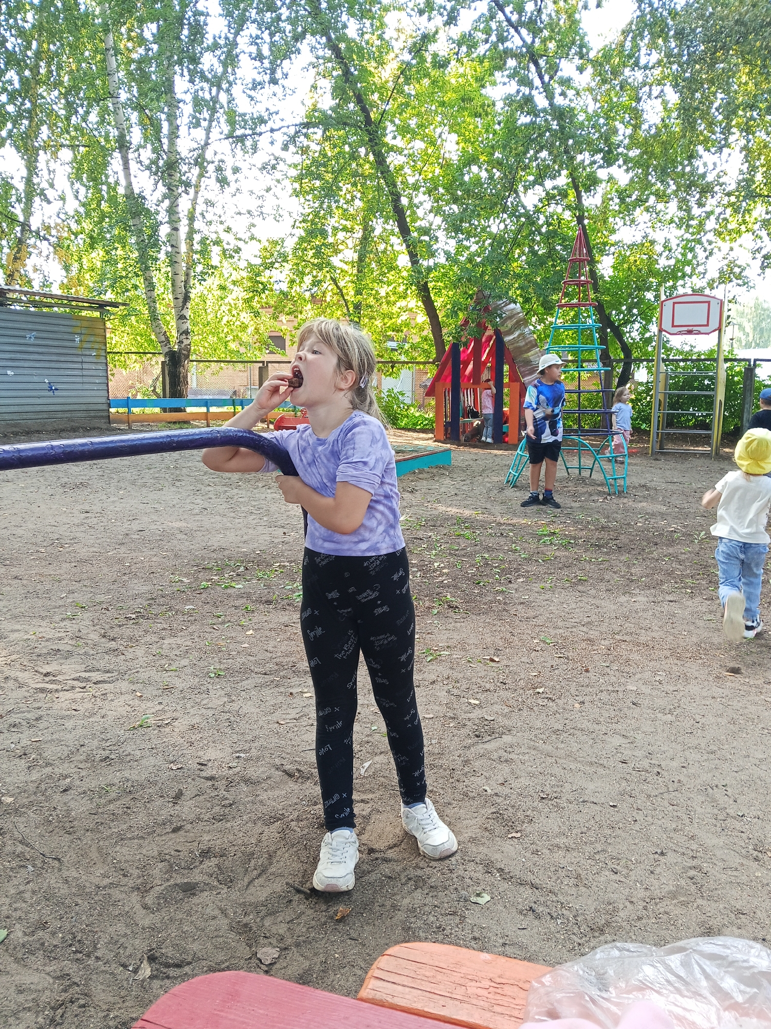 A young girl with blonde hair, wearing a purple t-shirt and black leggings with white text, stands in a sandy playground in Chaikovsky, Russia. She is holding a purple bar and putting a dark berry into her mouth. In the background, other children are playing on playground equipment, including a slide and a basketball hoop. Trees with green foliage surround the playground, and a fence is visible in the distance. The ground is covered in sand and scattered leaves.