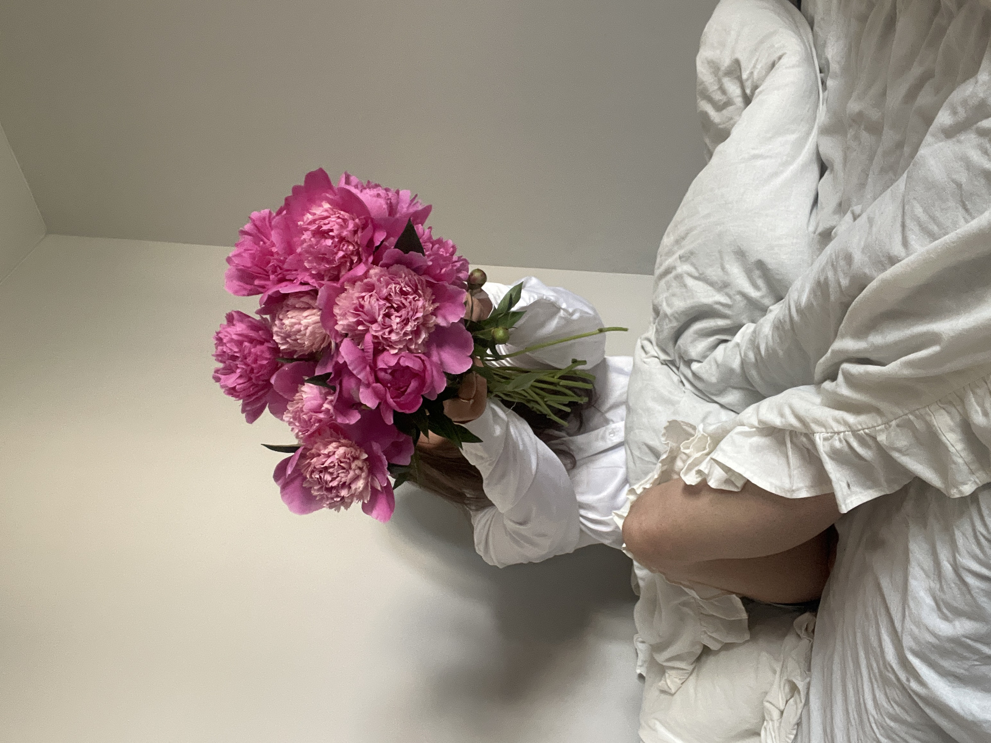 A person is seated on a bed, holding a bouquet of pink peonies in front of their face, obscuring their features. They are wearing a white collared shirt and their legs, clad in dark shorts, are visible beneath the ruffled white duvet. The bed is covered in rumpled white linens, and the background consists of a plain, light-colored wall. The scene is illuminated by soft, natural light. The specific location within Ternopil, Ukraine is not discernible from the image.