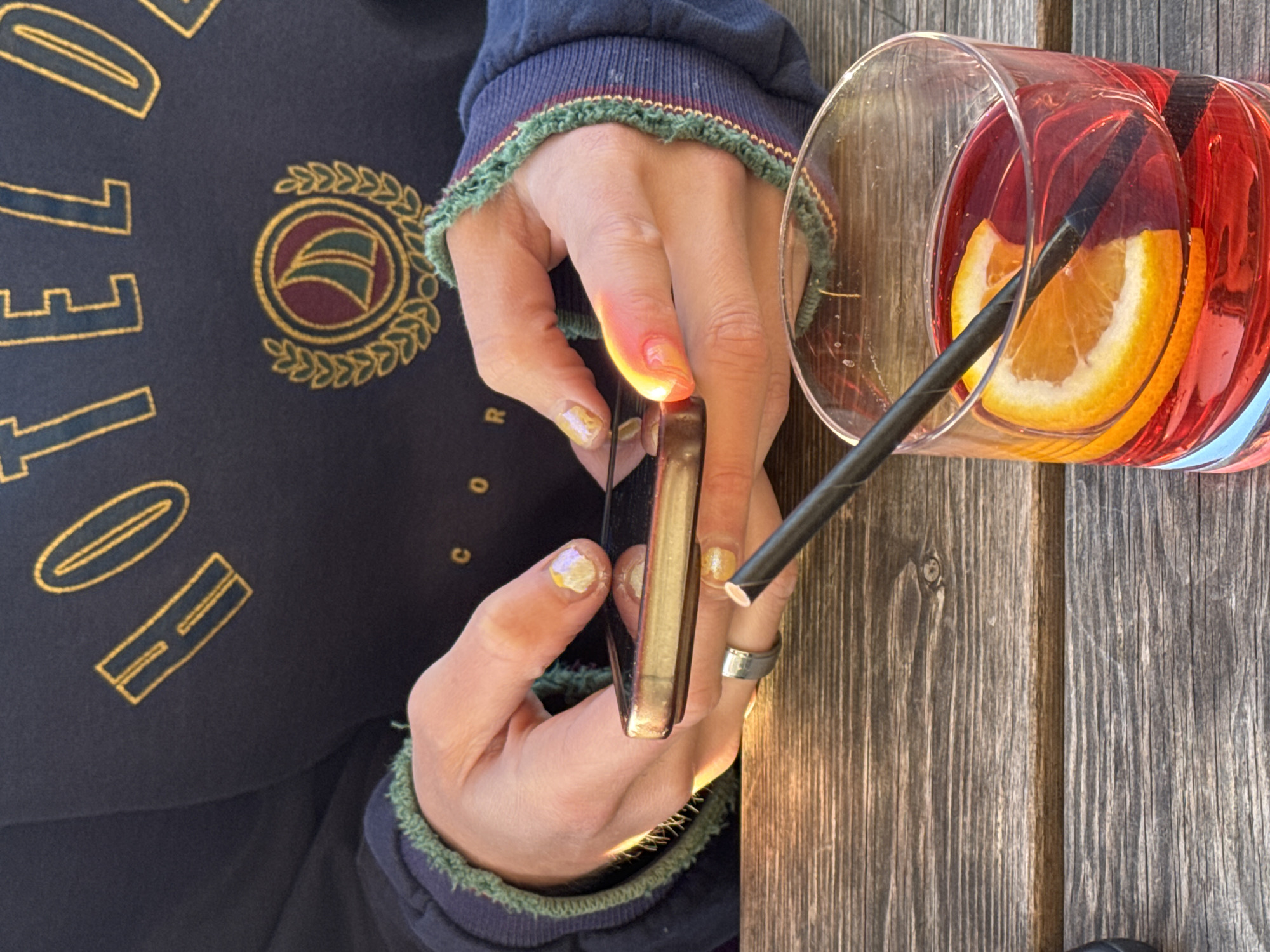 The image, taken from an overhead perspective, captures a person's hands holding a smartphone on a wooden table. The person is wearing a dark blue sweatshirt with embroidered gold text, partially revealing "HELLO" on the left and faint letters "C O R" to the right. A circular crest with a gold laurel wreath surrounds a maroon and green design on the sweatshirt. The cuffs of the sweatshirt are dark blue with a green and red striped trim.
The hands have shimmering, possibly gold or iridescent, nail polish. One finger wears a simple silver ring. The smartphone, held vertically, has a bright glow reflecting off its edge and the person's finger, suggesting either strong sunlight hitting the screen or a light source from the phone itself.
Beside the phone, a glass filled with a reddish-orange drink is visible, containing a slice of orange and a black paper straw. The beverage appears refreshing, possibly a cocktail or spritzer. The table is made of rustic, weathered wooden planks. The bright, natural lighting indicates it is daytime, likely sunny, given the strong highlights and reflections. The overall scene suggests a casual outdoor setting, such as a cafe, patio, or backyard in London, United Kingdom.