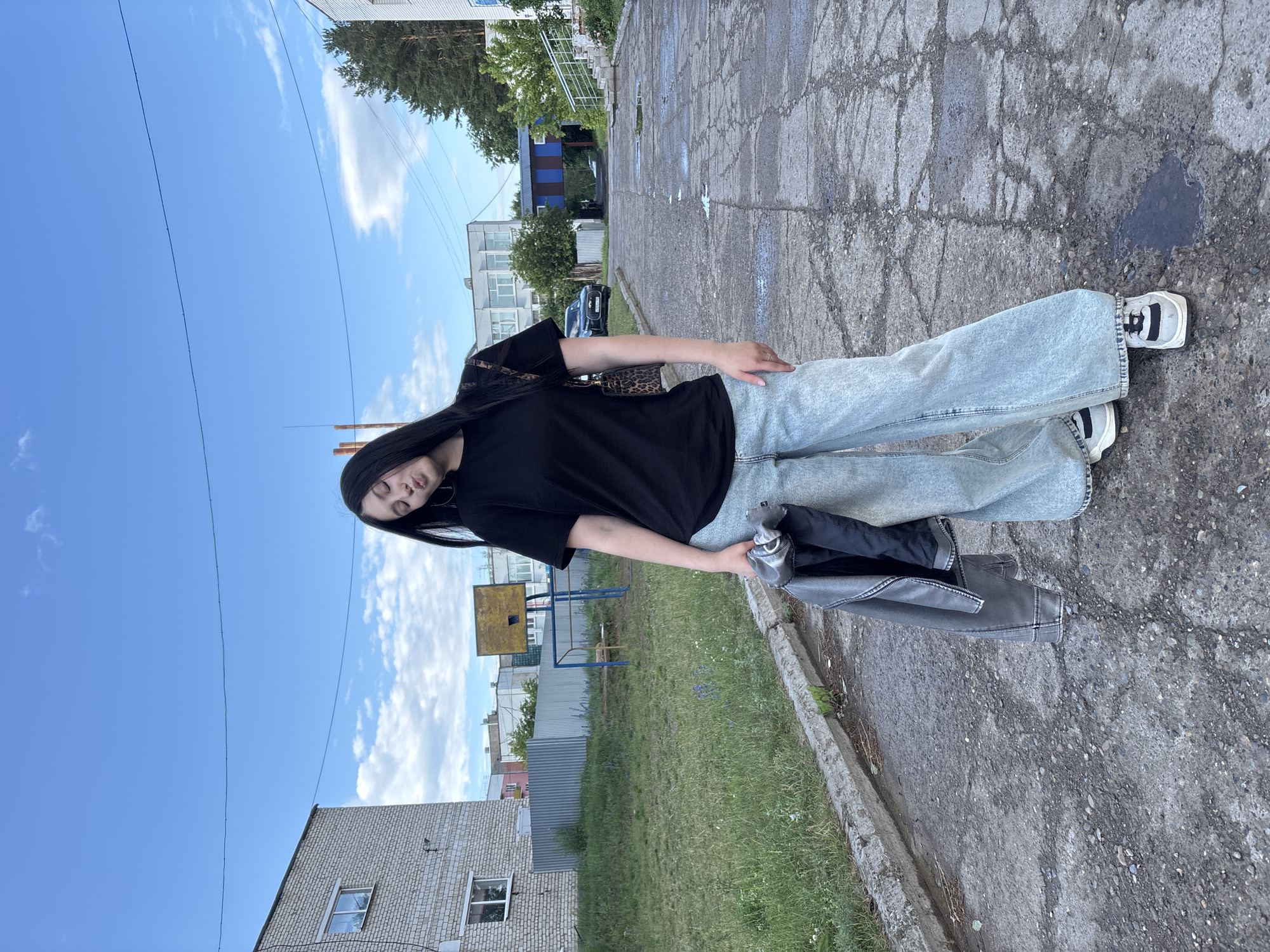 A young woman with long dark hair stands in front of a building, holding a grey jacket in her hand. She is wearing a black t-shirt, light blue wide-leg jeans, and white sneakers with black accents. Her body is angled towards the camera, and she appears to be looking away with her eyes closed, possibly posing. A patterned purse with a leopard print strap is visible over her shoulder. The scene is outdoors, with a cracked asphalt surface in the foreground and a grassy area with a basketball hoop and chain-link fence behind her. Several buildings and trees are visible in the background, under a bright blue sky with scattered white clouds. The lighting suggests it is daytime, likely afternoon, and the weather appears fair. There is no visible text in the image. The overall setting suggests an urban or semi-urban environment in Chaikovsky, Russia.